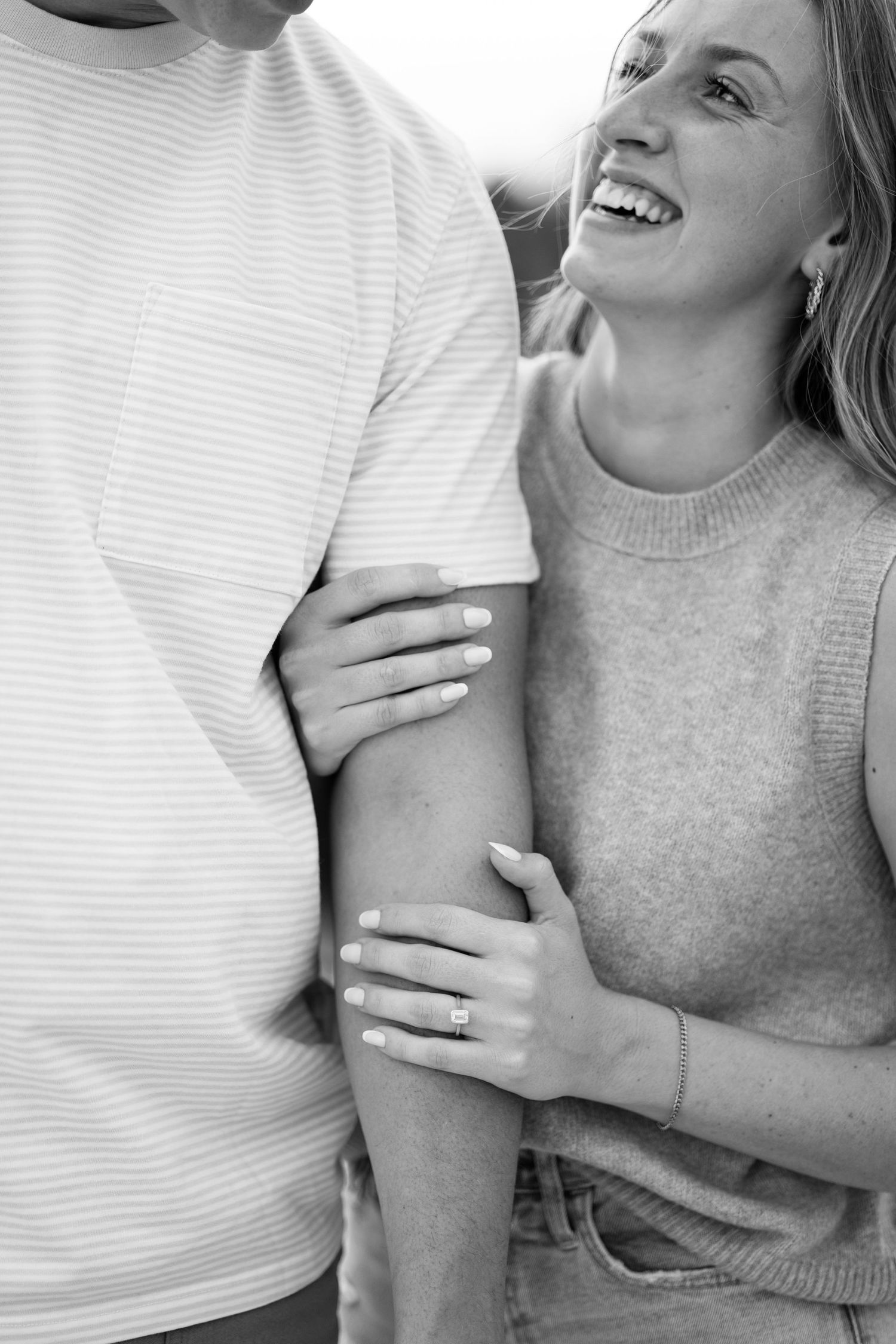 Black and white close-up of smiling woman embracing mans arm, showing engagement ring.
