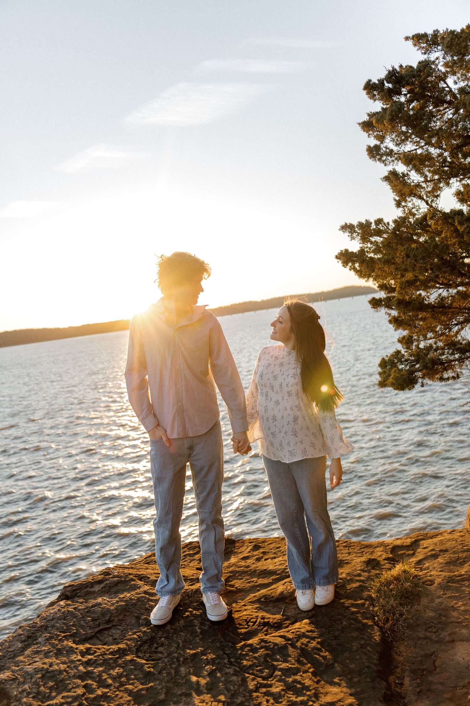 Couple holding hands on rocky shoreline at golden sunset with glowing water and pine tree backdrop.