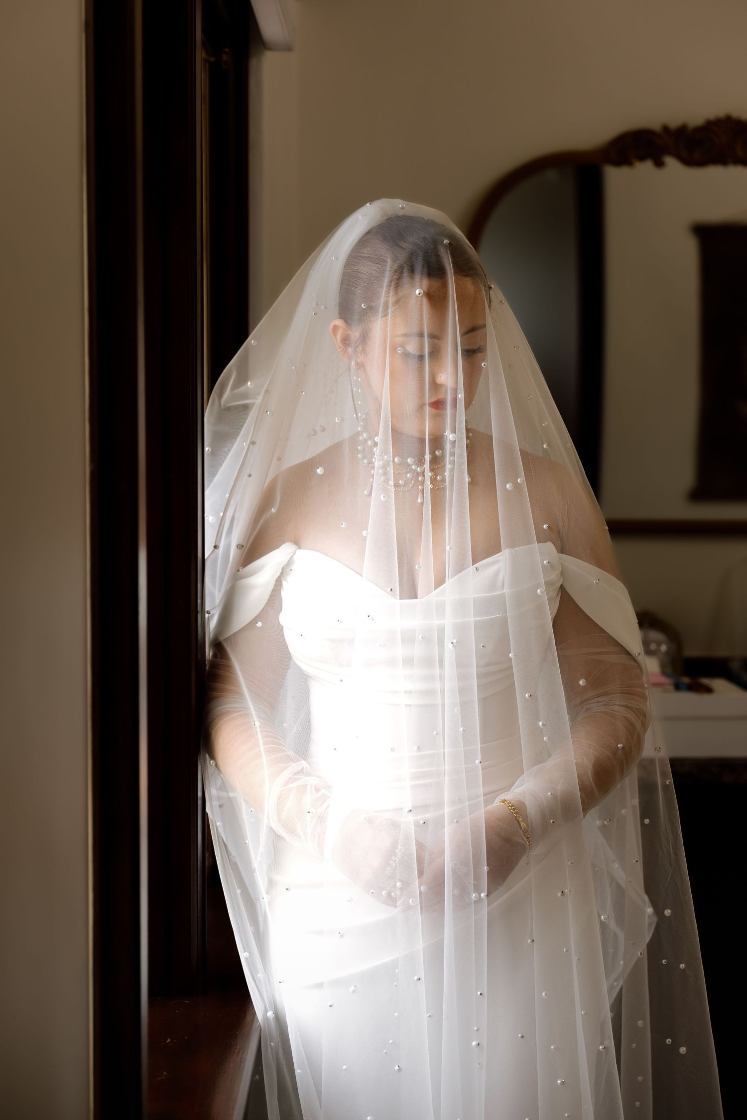Bride in white strapless gown with pearl-adorned veil covering her face in soft indoor light.