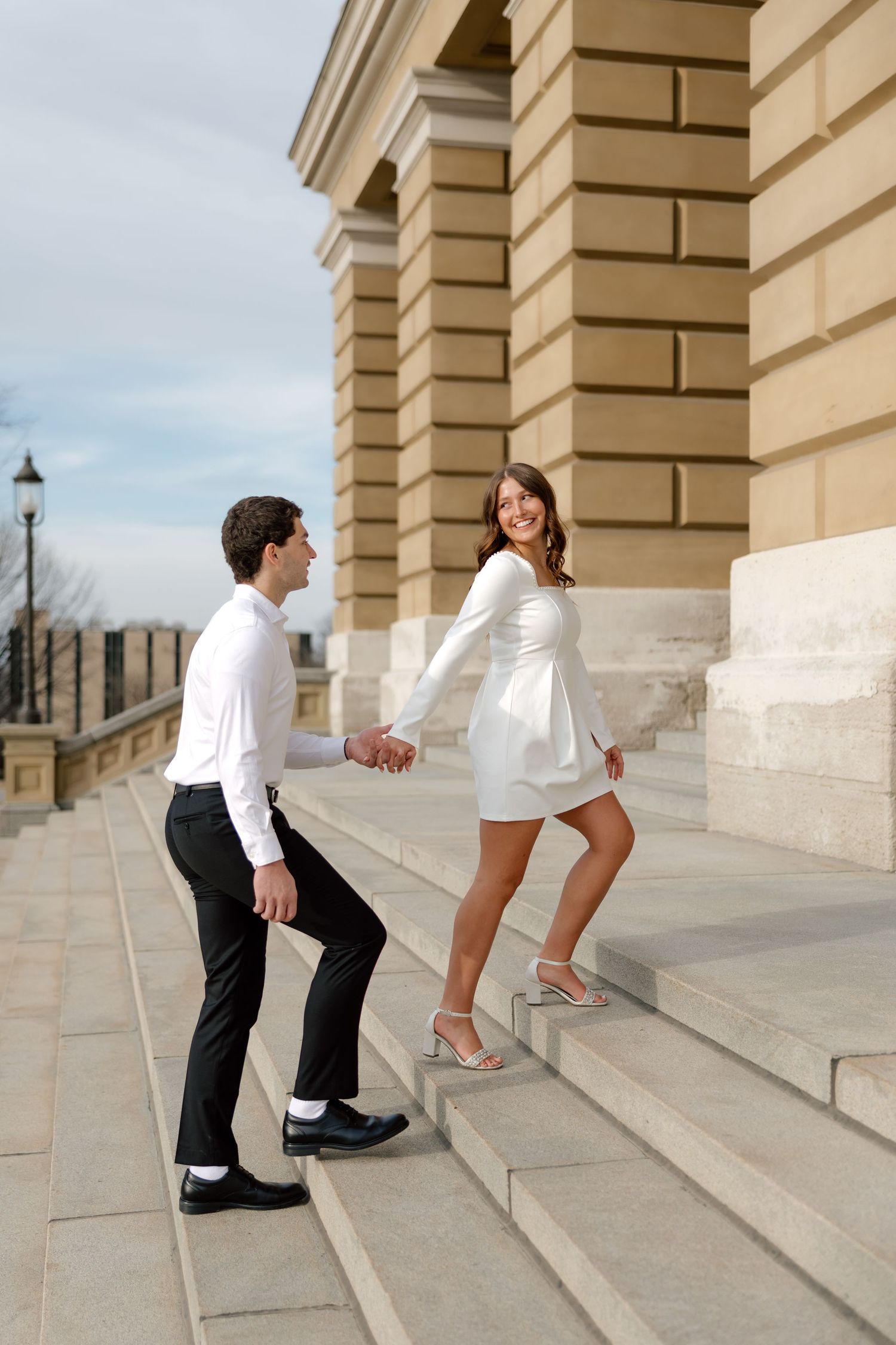 Smiling bride in white mini dress leads groom up stone steps outside a classical building.