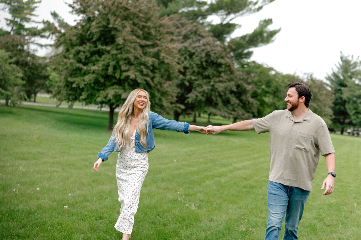 A smiling couple holds hands while walking in a lush green park, the woman leading in a floral dress and denim jacket.