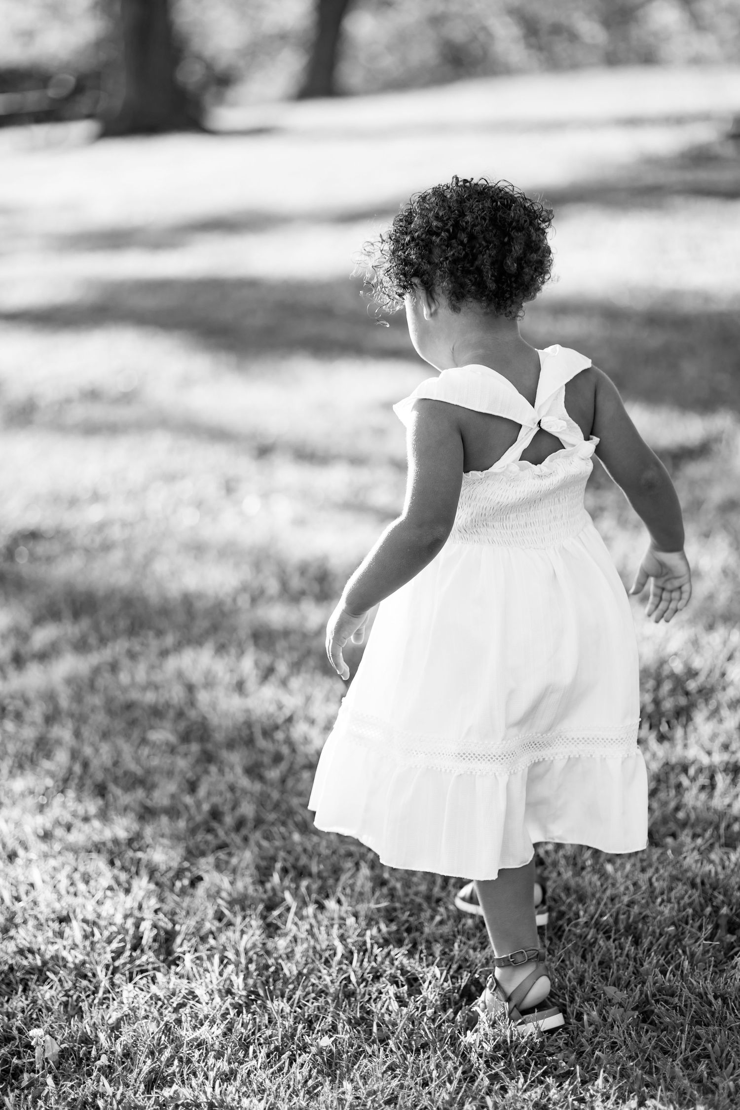 Black and white photo of a young girl in a white dress walking away on green grass in a park.