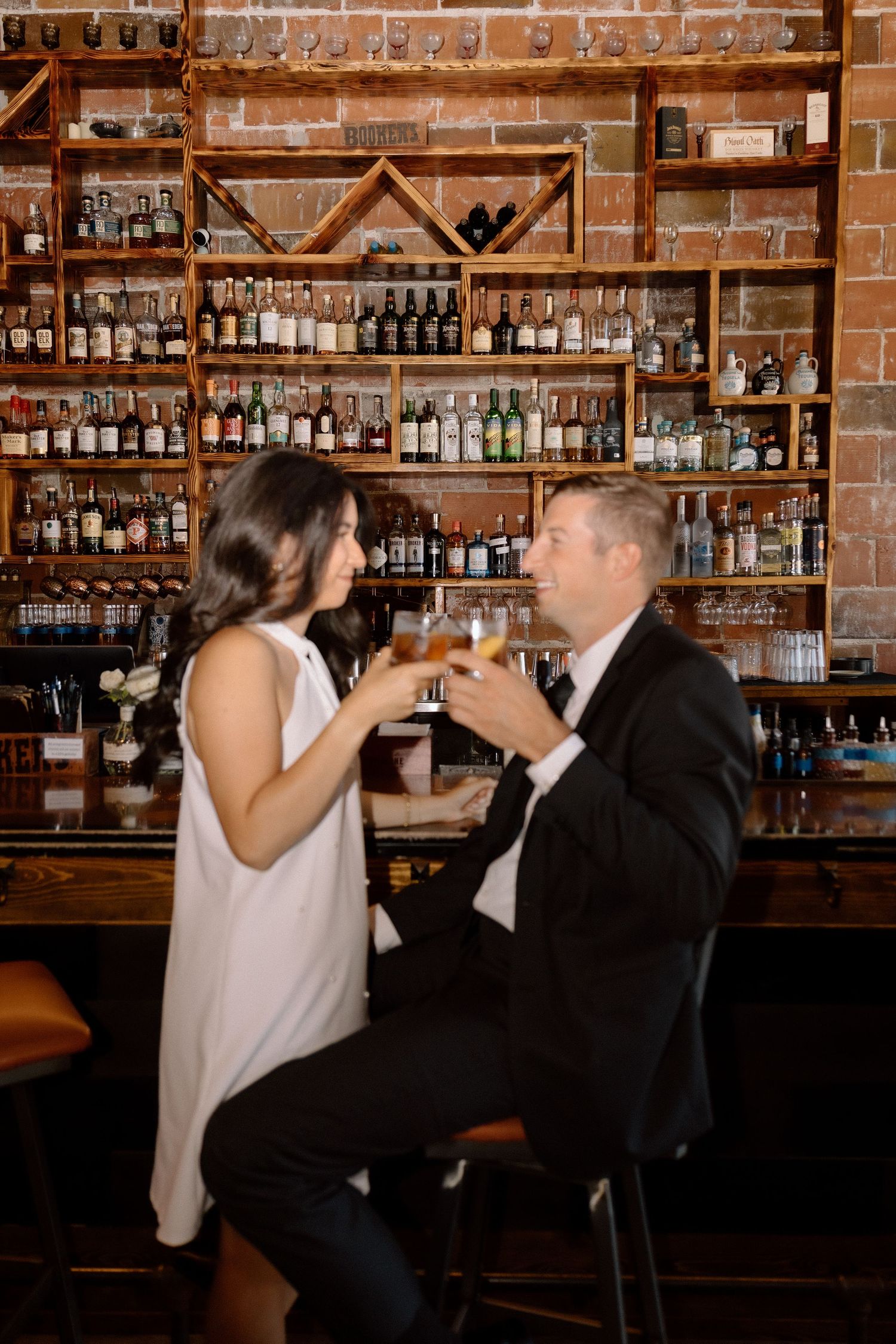 Couple toasting cocktails at a rustic bar with brick walls and shelves of bottles behind them.