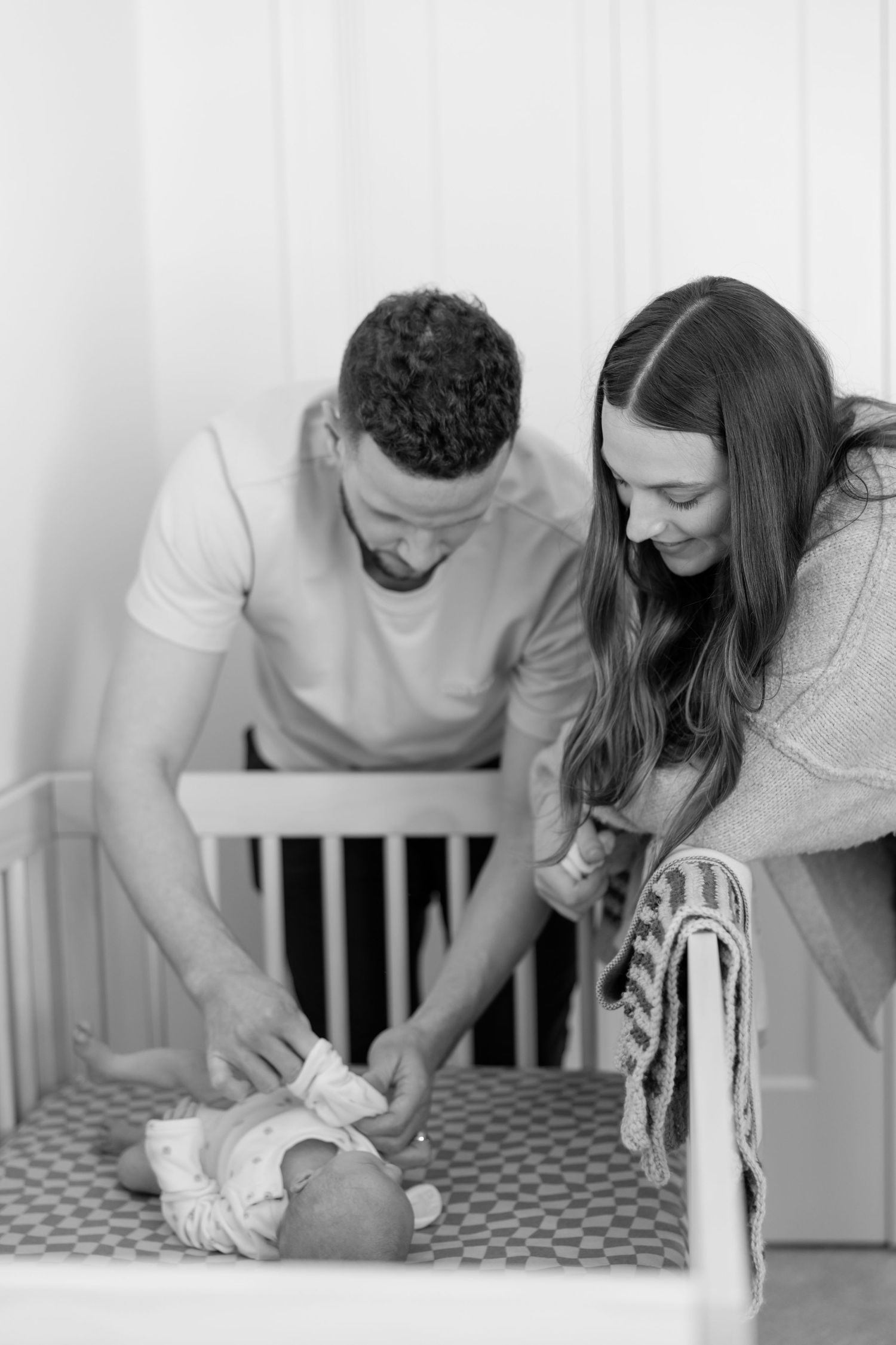 Black and white photo of parents lovingly tending to their newborn baby in a crib.