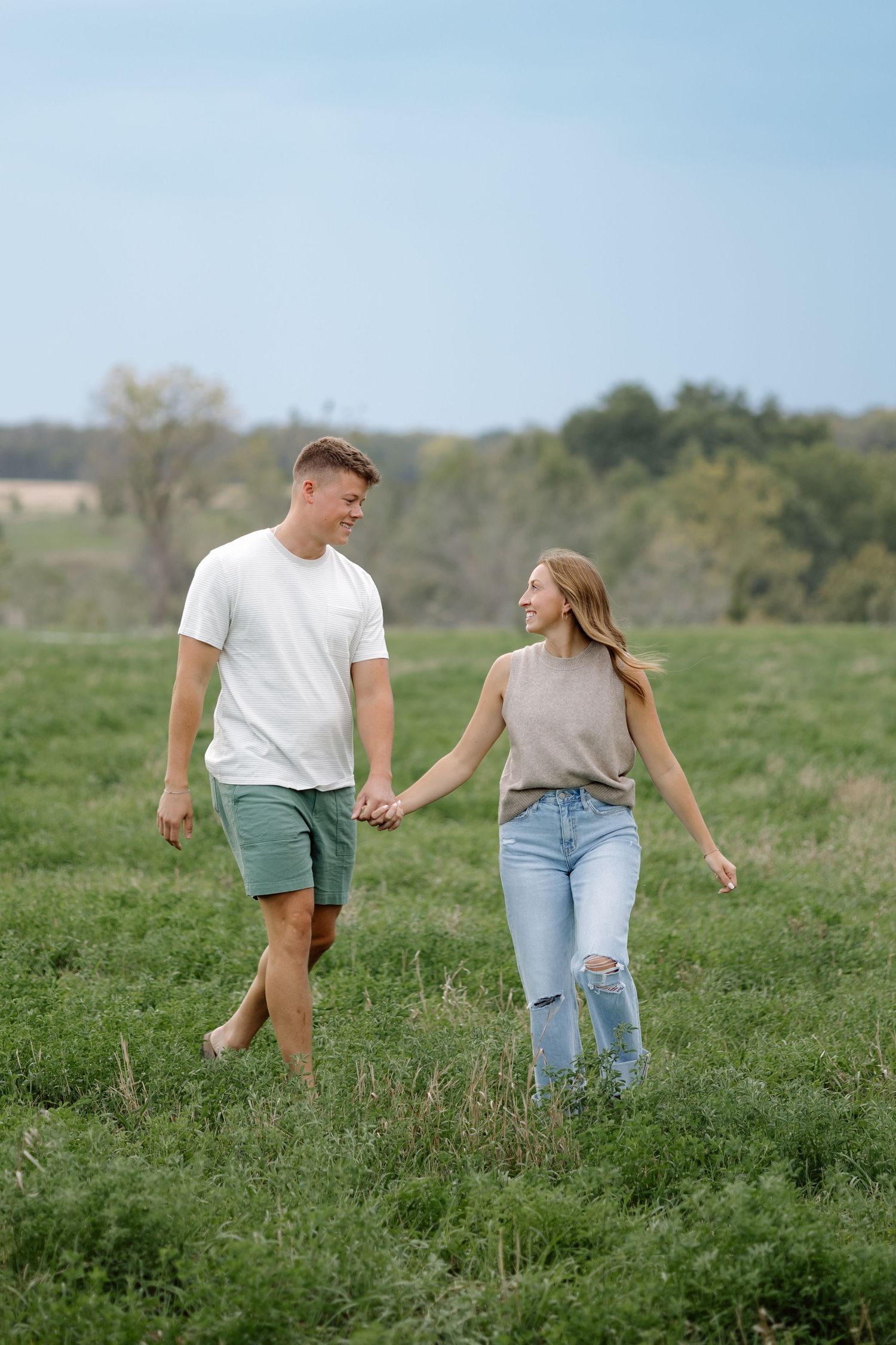 A happy couple holds hands while walking through a lush green field on a sunny day.