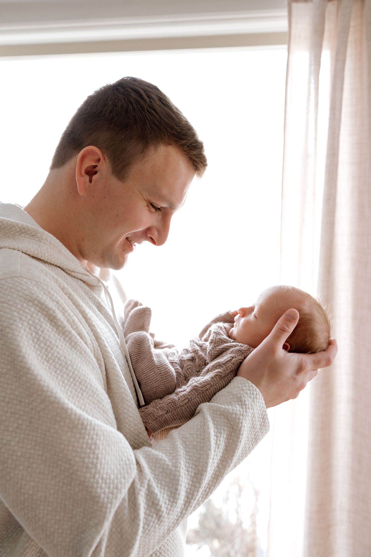 Proud father in white textured shirt smiles while carefully holding his newborn baby by a window.