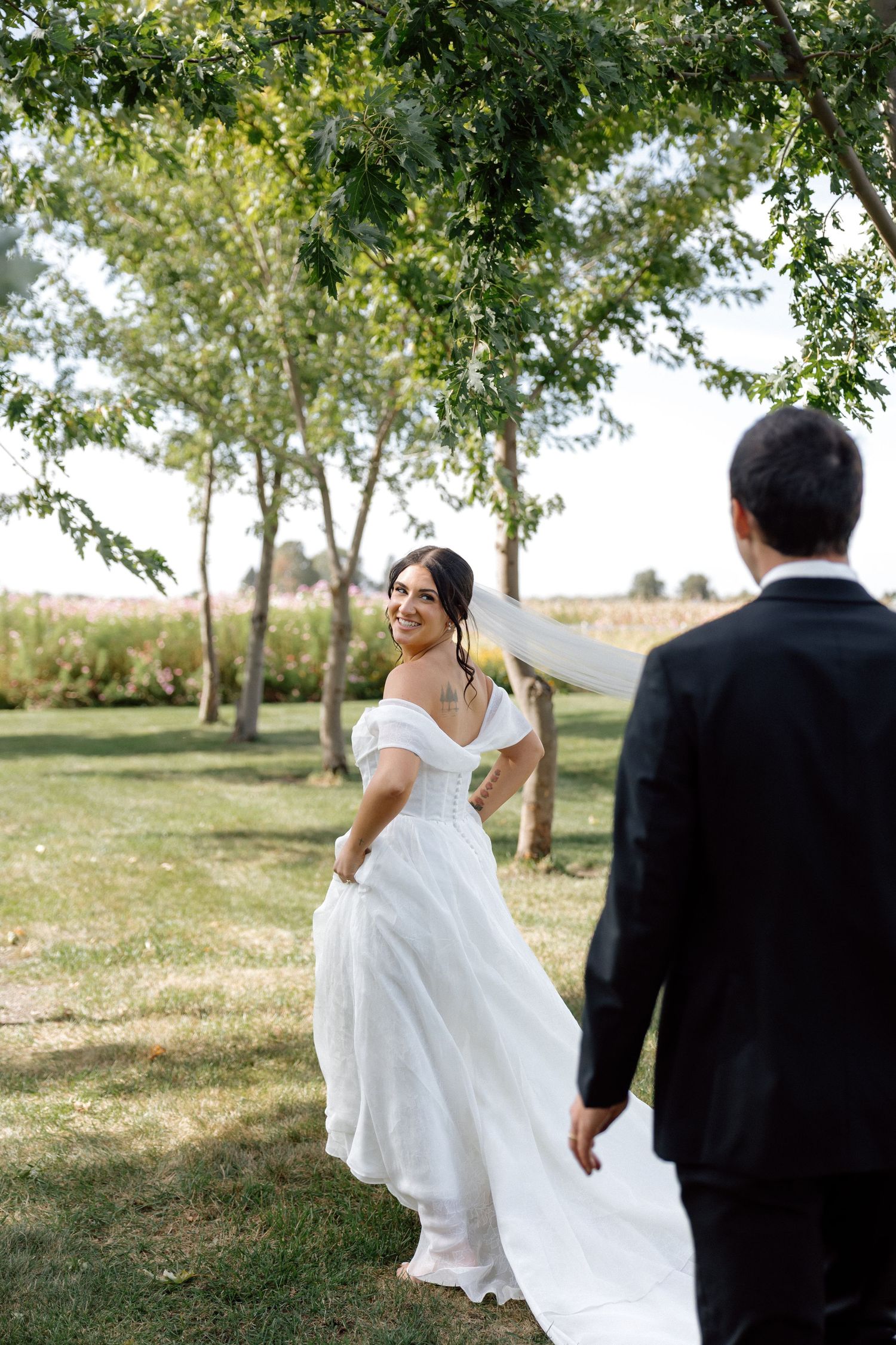 Smiling bride in white off-shoulder gown twirling toward groom in tree-lined garden.