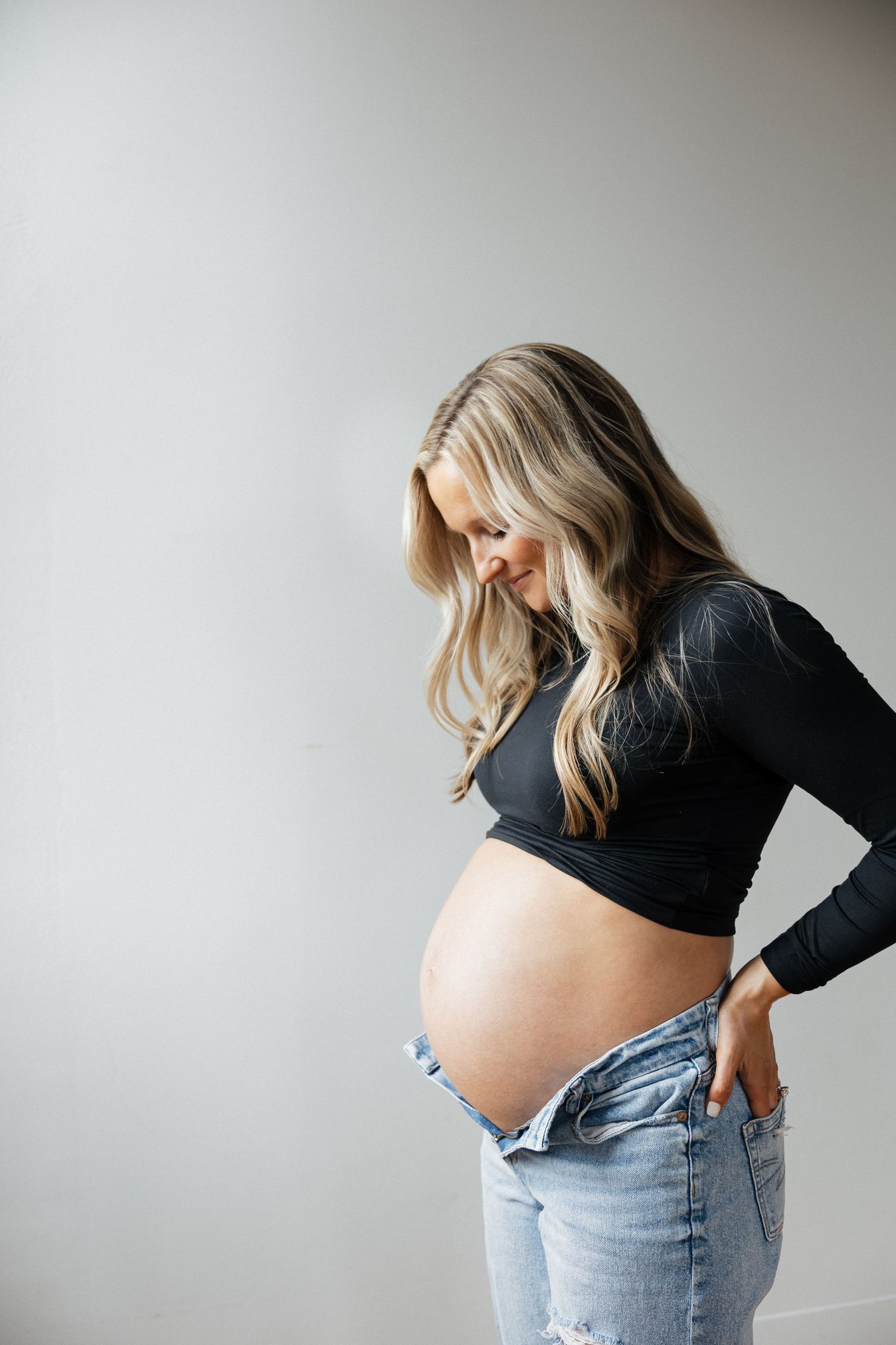 Pregnant woman in black long-sleeve crop top gazing down at her bare baby bump with ripped jeans lowered.