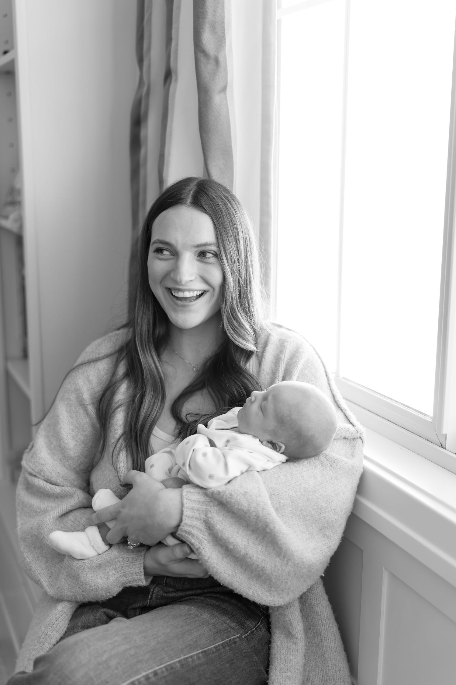 Black and white portrait of a laughing mother cradling her newborn by a window.