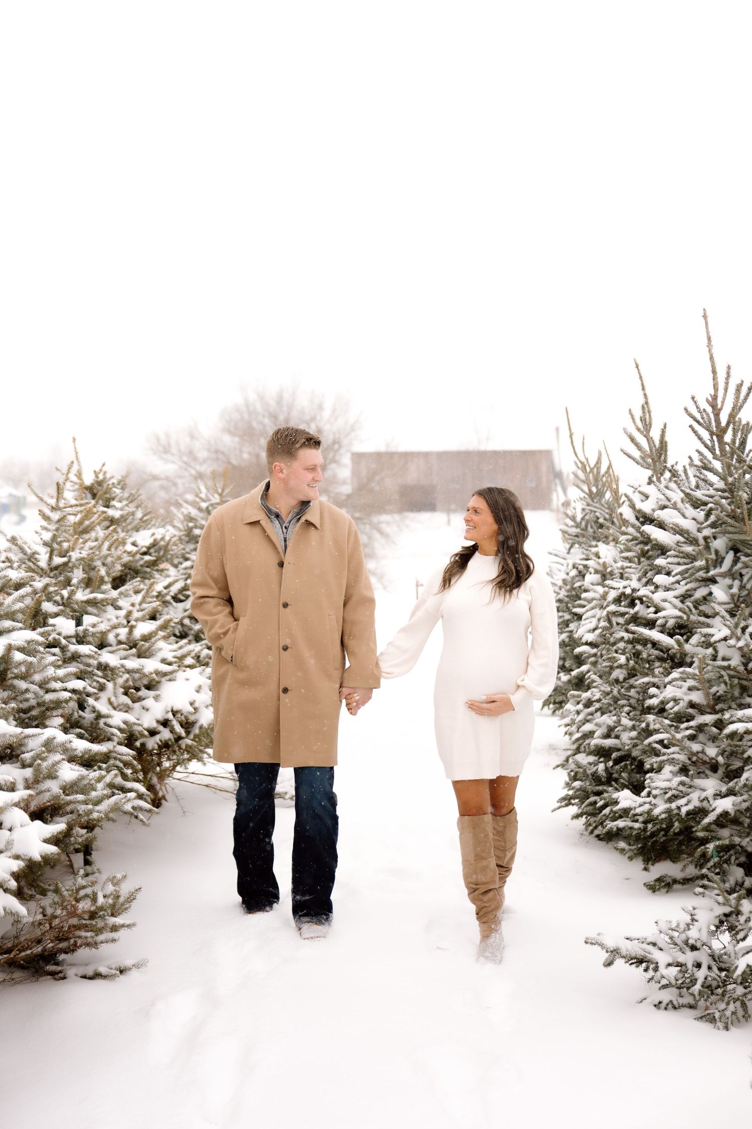 A pregnant couple walks hand-in-hand through a snowy Christmas tree farm, smiling at each other.