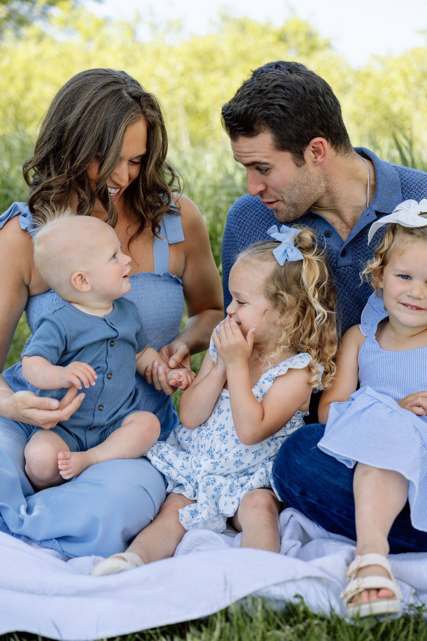 Family of five sitting on blanket outdoors in coordinating blue outfits, parents smiling at young children.