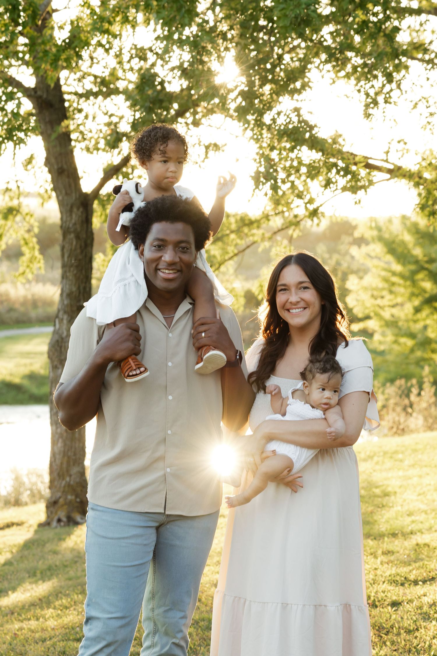 Smiling interracial family posing outdoors at sunset, father holding toddler on shoulders, mother cradling newborn.