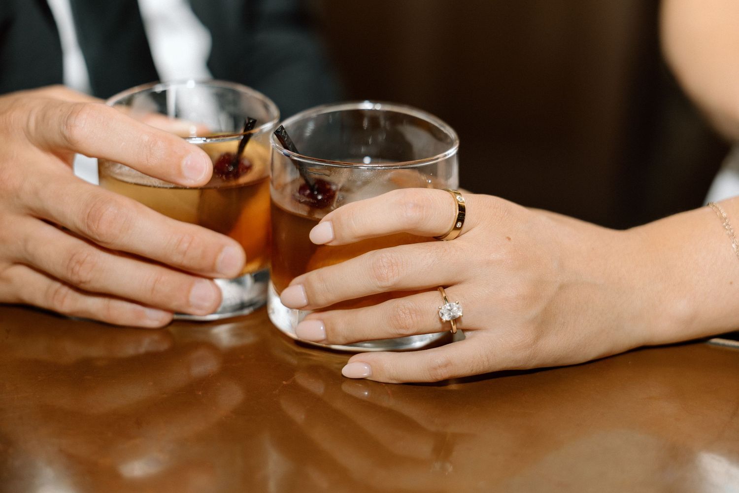 Close-up of couple clinking whiskey glasses on a bar, woman wearing a diamond engagement ring.