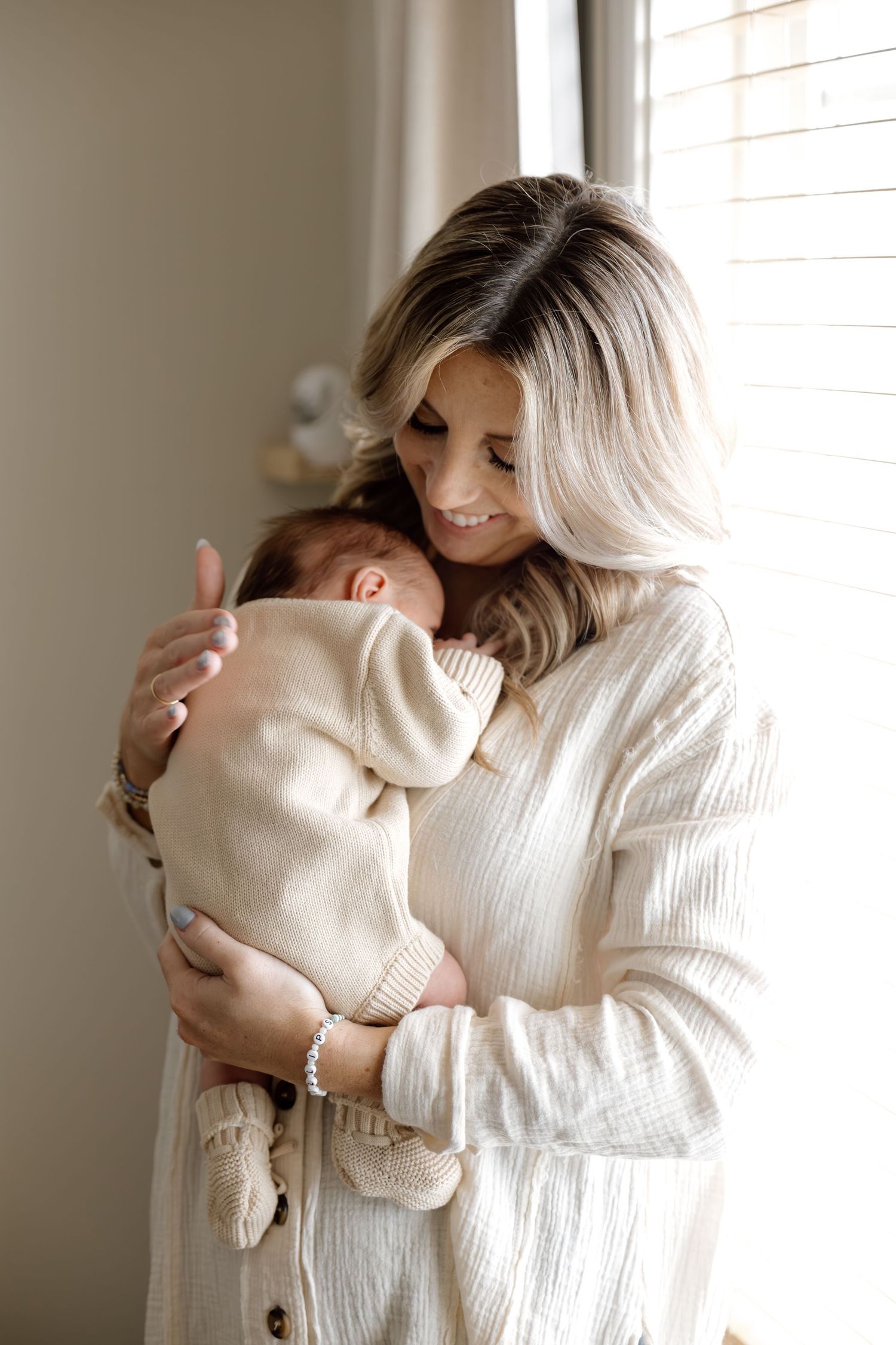 Smiling mother in cream sweater tenderly holding sleeping newborn by window.