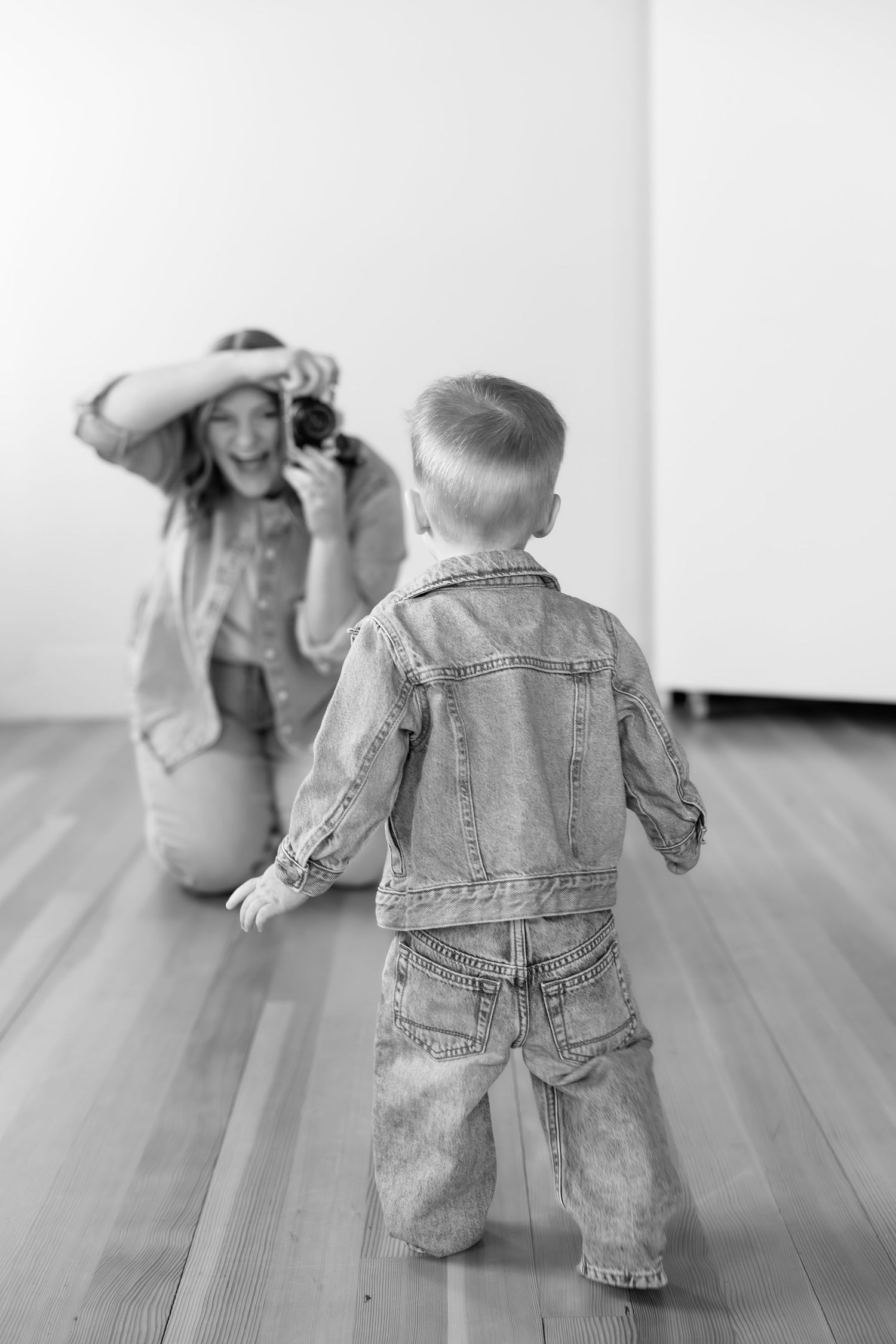 Black and white photo of a photographer capturing a toddler boy walking toward her in a studio.