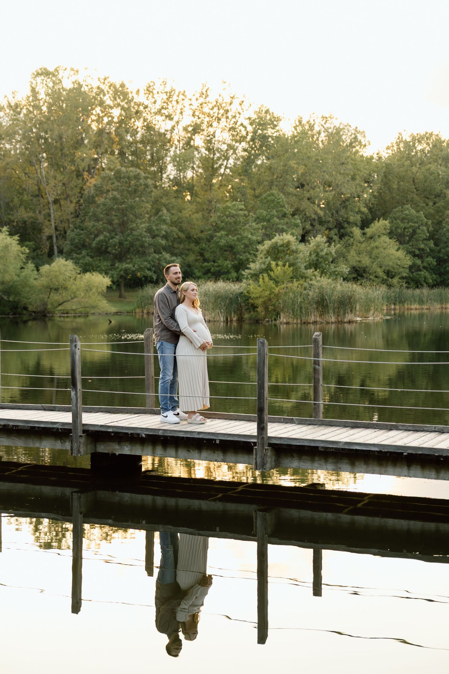 Couple embracing on lakeside dock at golden hour with their reflection visible in water.