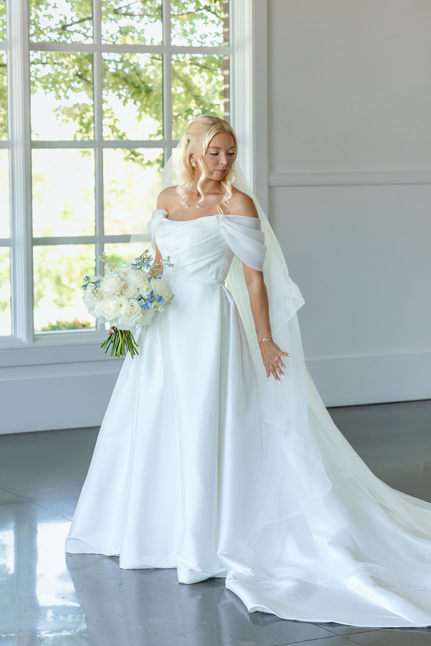 Elegant bride in an off-shoulder white satin ball gown holding a white and blue bouquet by a large window.