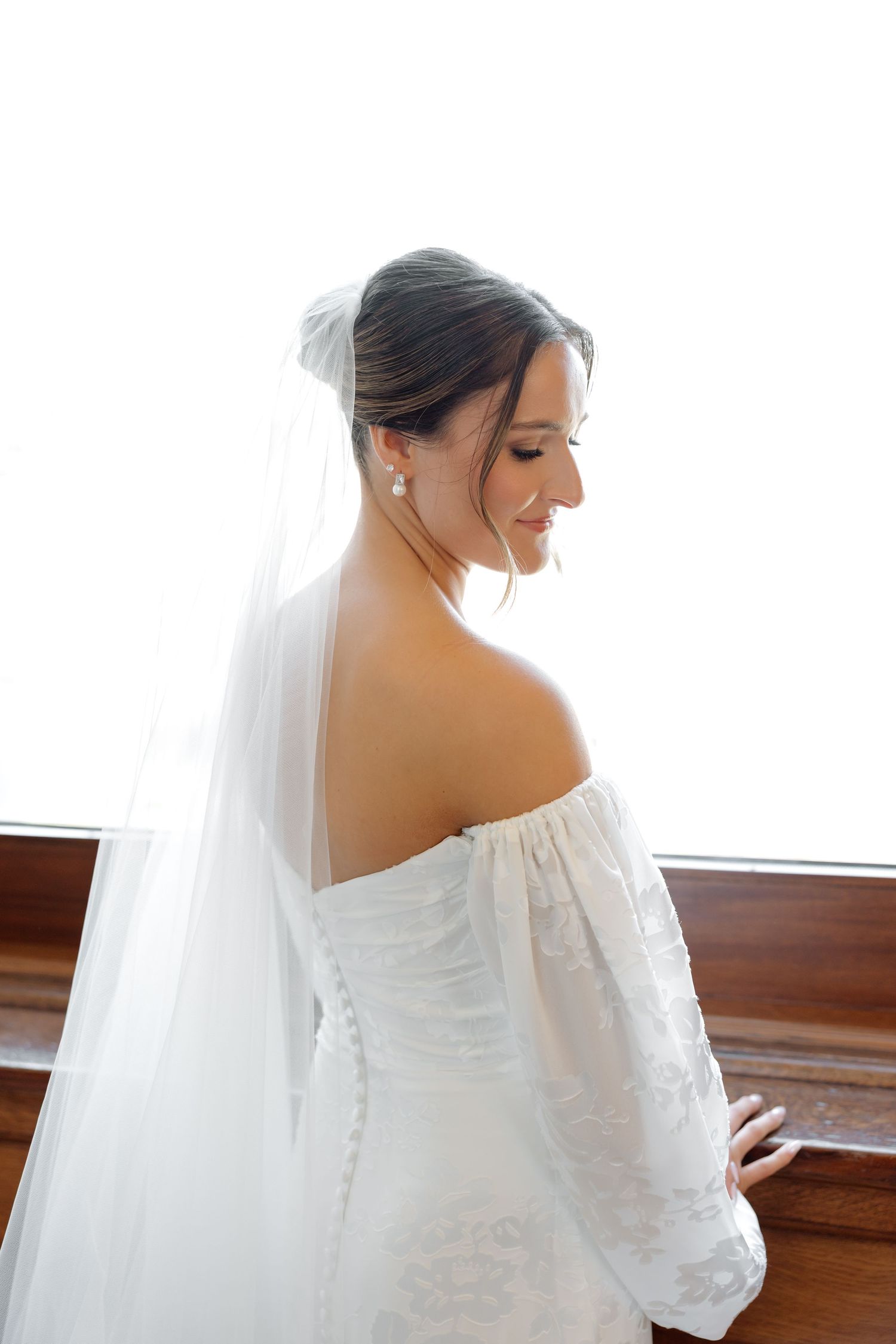 Elegant bride in off-shoulder lace wedding dress with veil smiles softly near bright window.