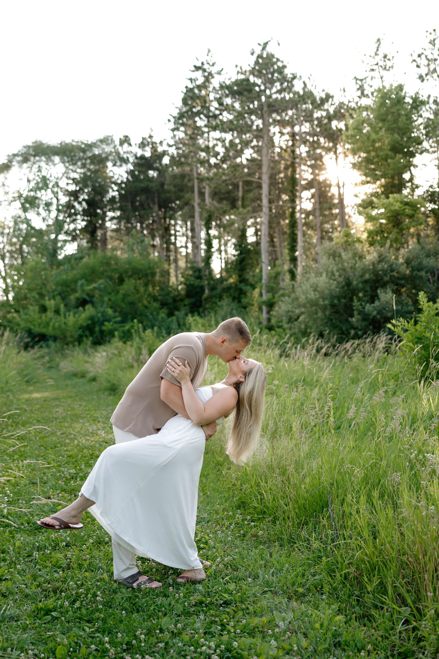 Couple sharing a romantic dip kiss in a lush green meadow with tall pine trees and golden sunset light behind them.