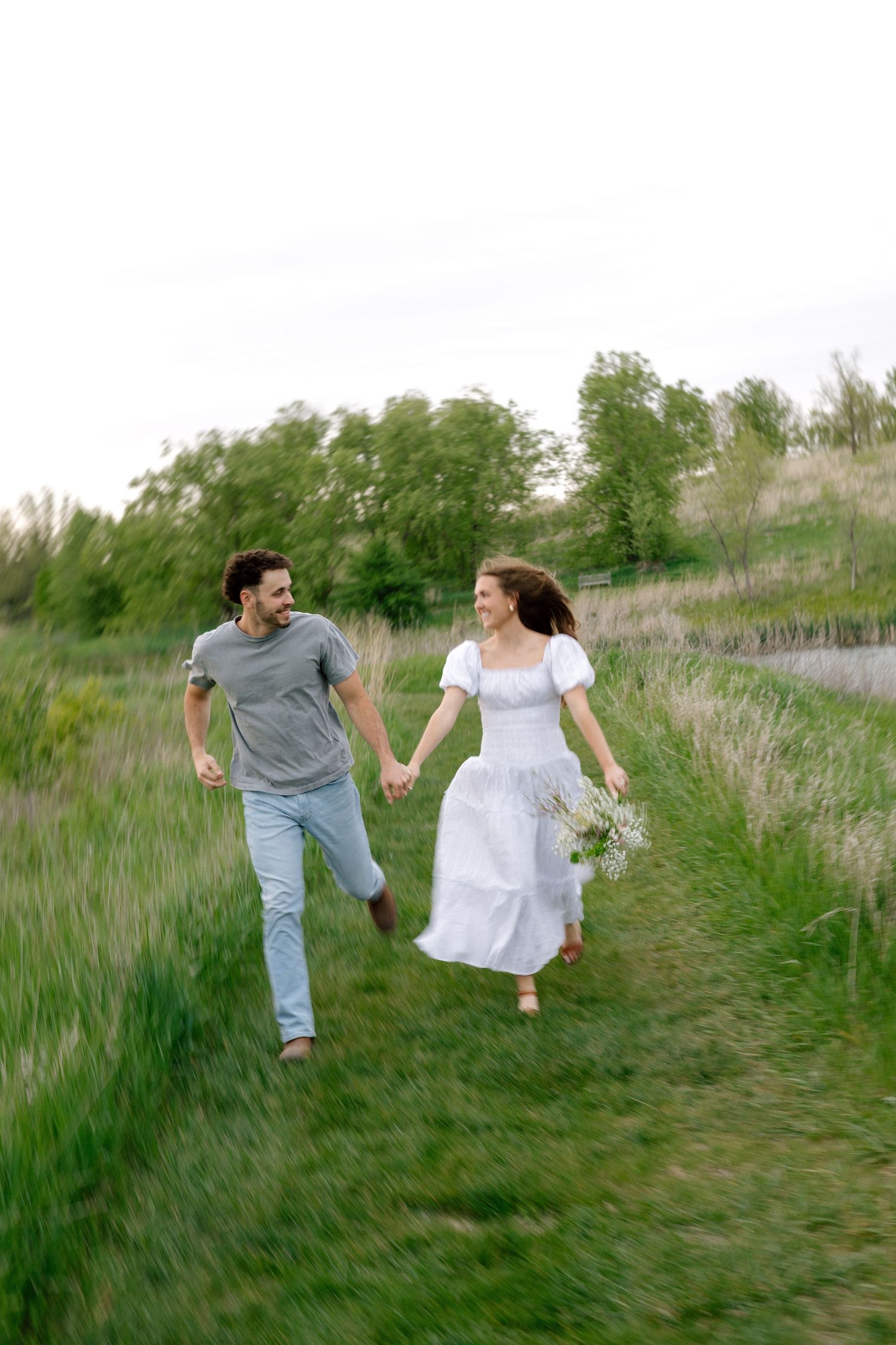 Happy couple holding hands running through green meadow, woman in white dress holding wildflowers, outdoor romantic photo.