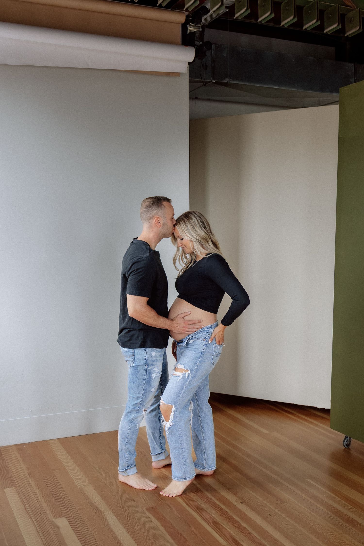 Expecting couple standing together in a photography studio, tenderly touching the pregnant woman's baby bump.