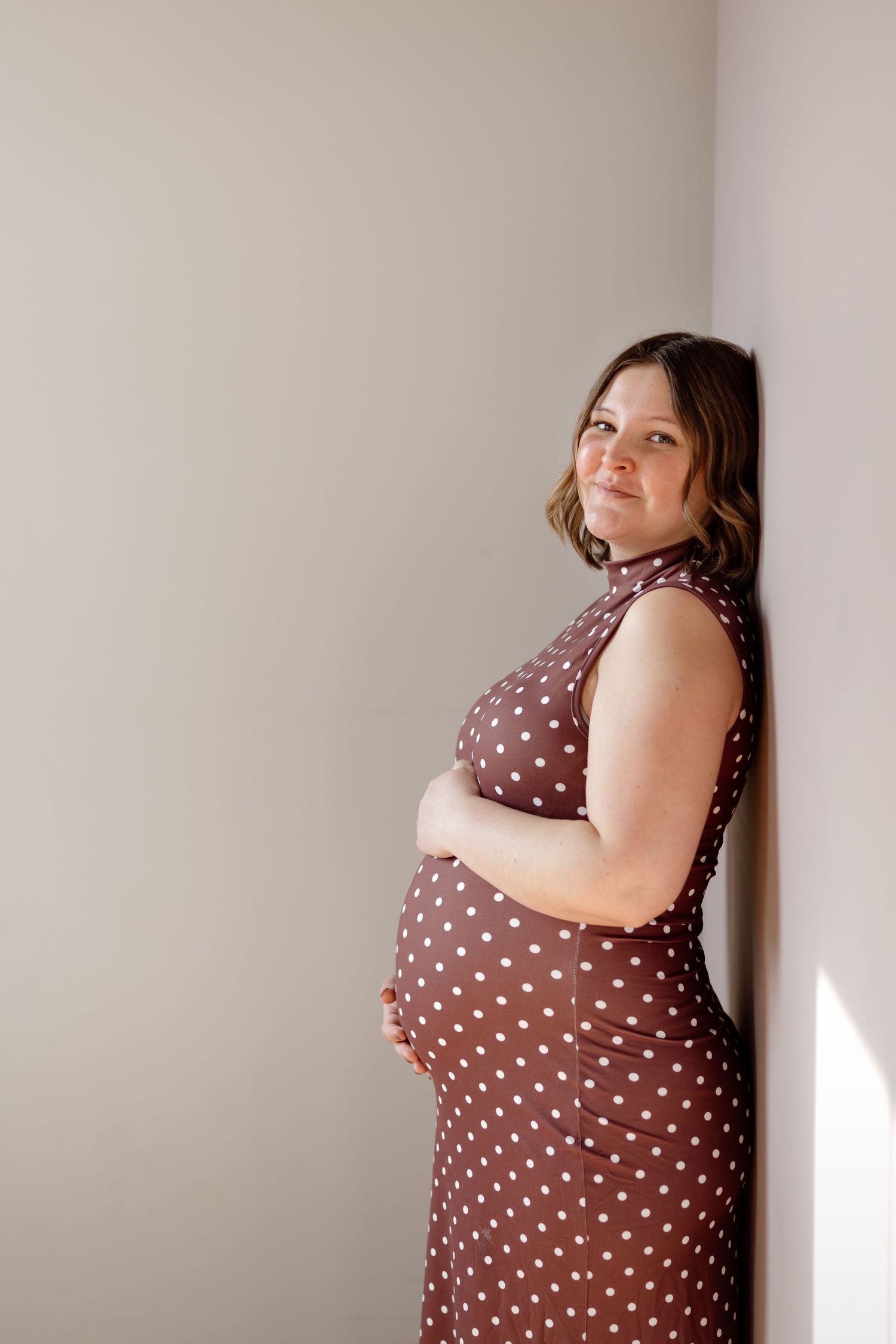 A pregnant woman in a brown polka dot dress leans against a wall, smiling softly while cradling her bump.