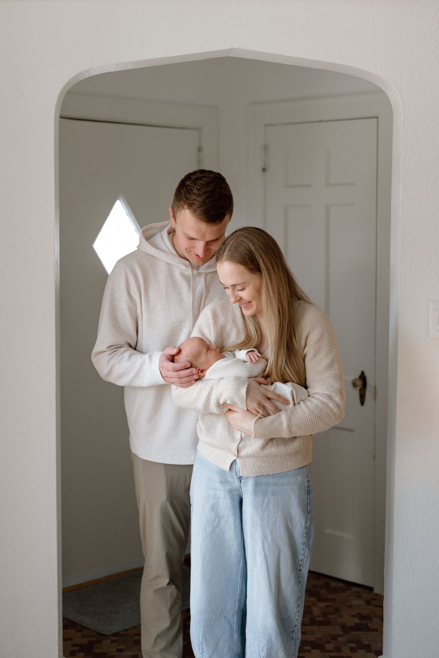 New parents stand together in an arched doorway, smiling down at their newborn baby in a bright white home.