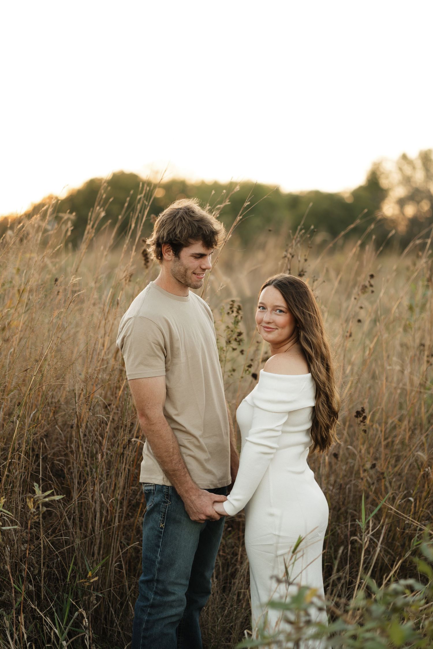 Couple holding hands in a golden sunlit field, woman in white dress and man in beige tee and jeans.