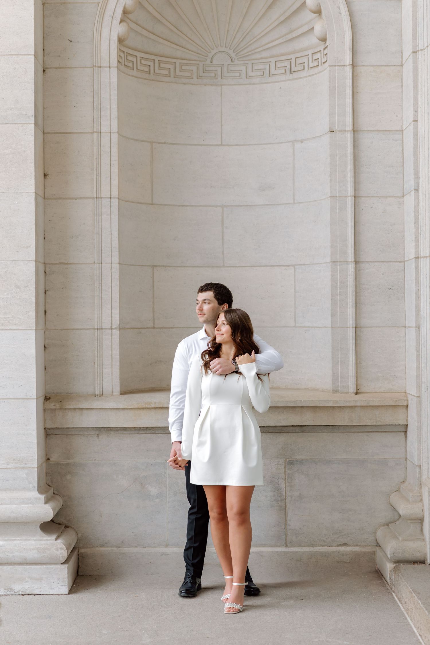 Couple embracing in front of ornate stone archway, woman in white mini dress.