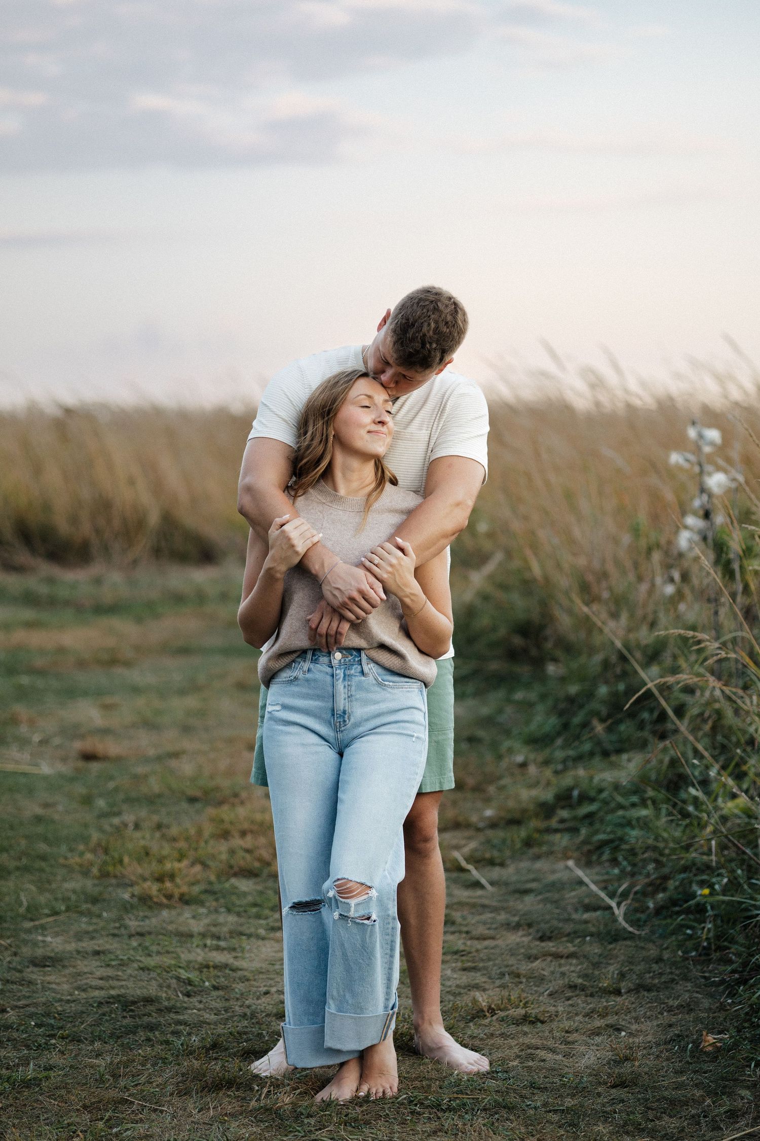 Man tenderly embracing woman from behind in golden grass field at sunset, both barefoot.