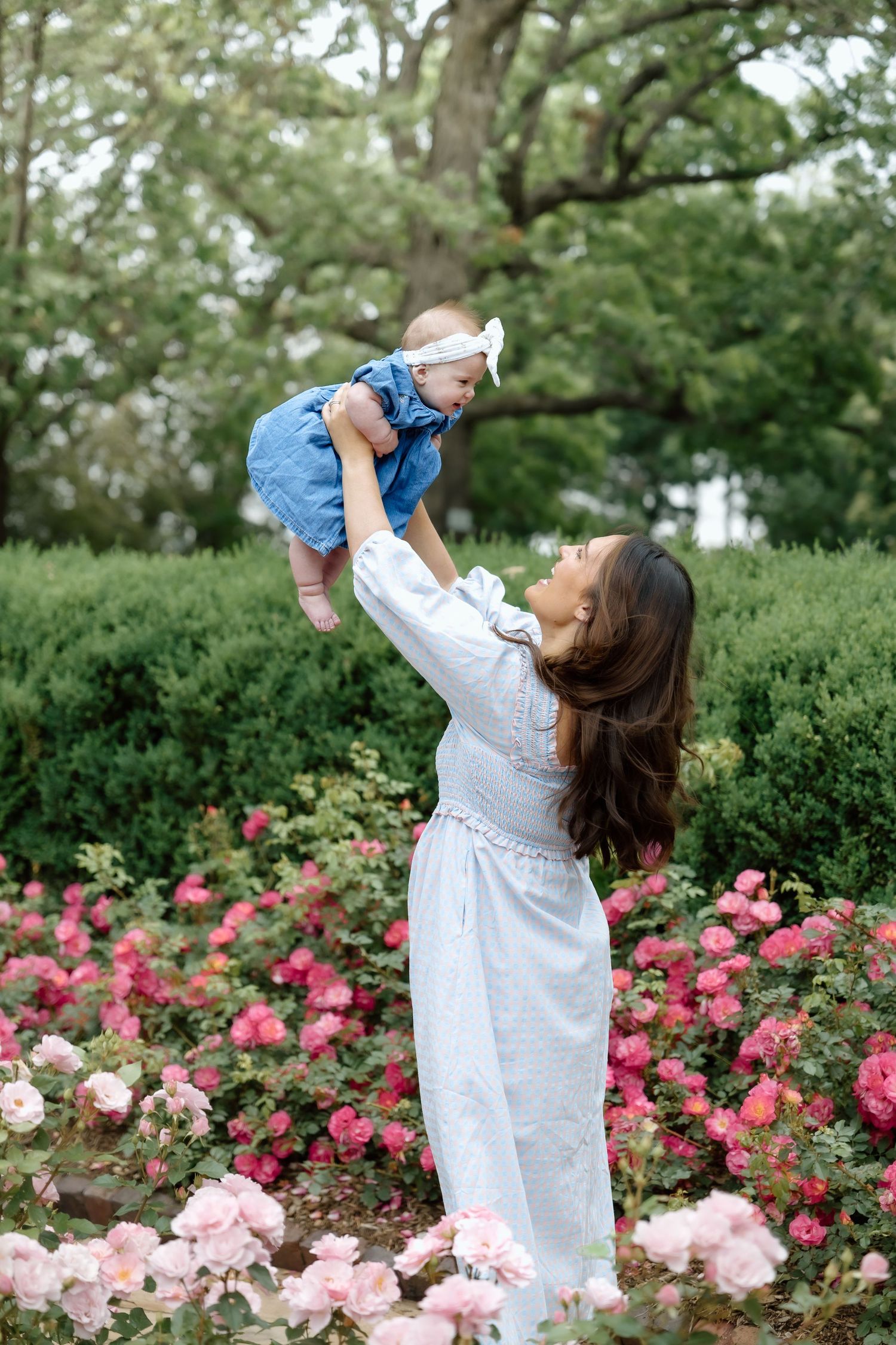 Mother in white dress lifting baby girl in blue dress among pink roses in a lush garden.