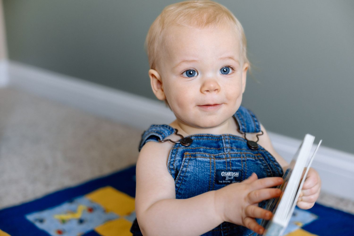 Adorable blue-eyed baby in denim overalls holding a book, sitting on a colorful play mat.