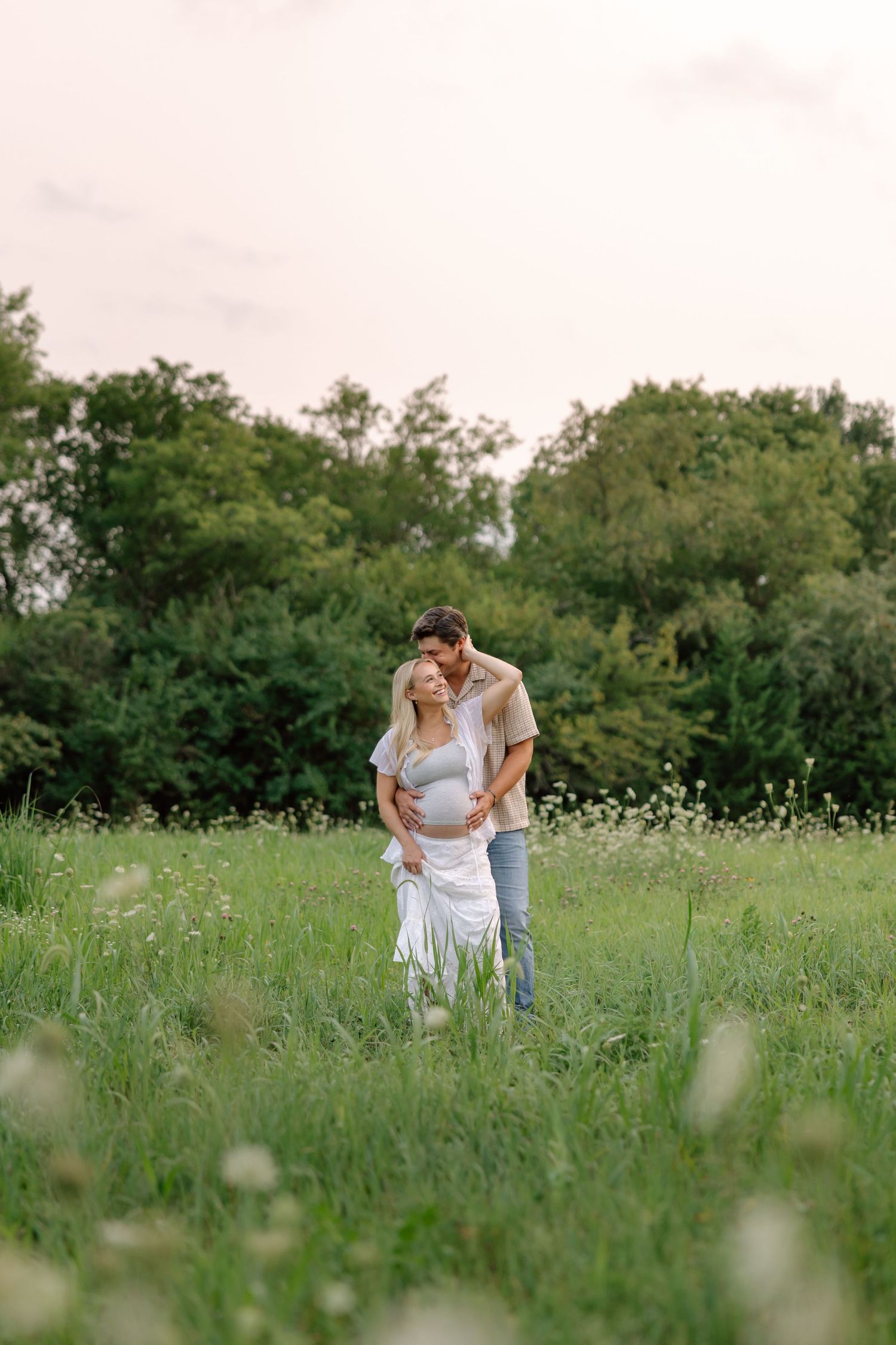 A couple stands in a lush green meadow, man embracing pregnant woman from behind, surrounded by wildflowers and trees.