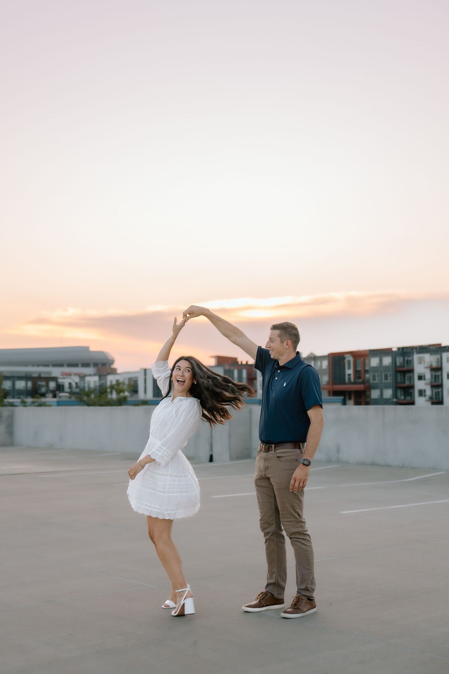 Man spinning his laughing partner on a rooftop parking garage during a beautiful pastel sunset.