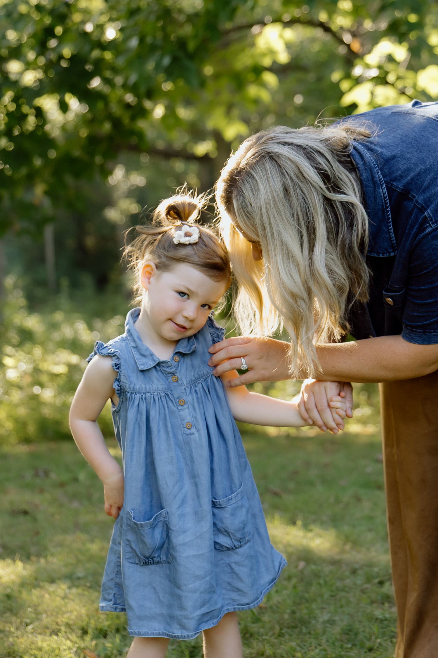 Blonde mother bending down to kiss young daughter in blue denim dress in sunny garden.