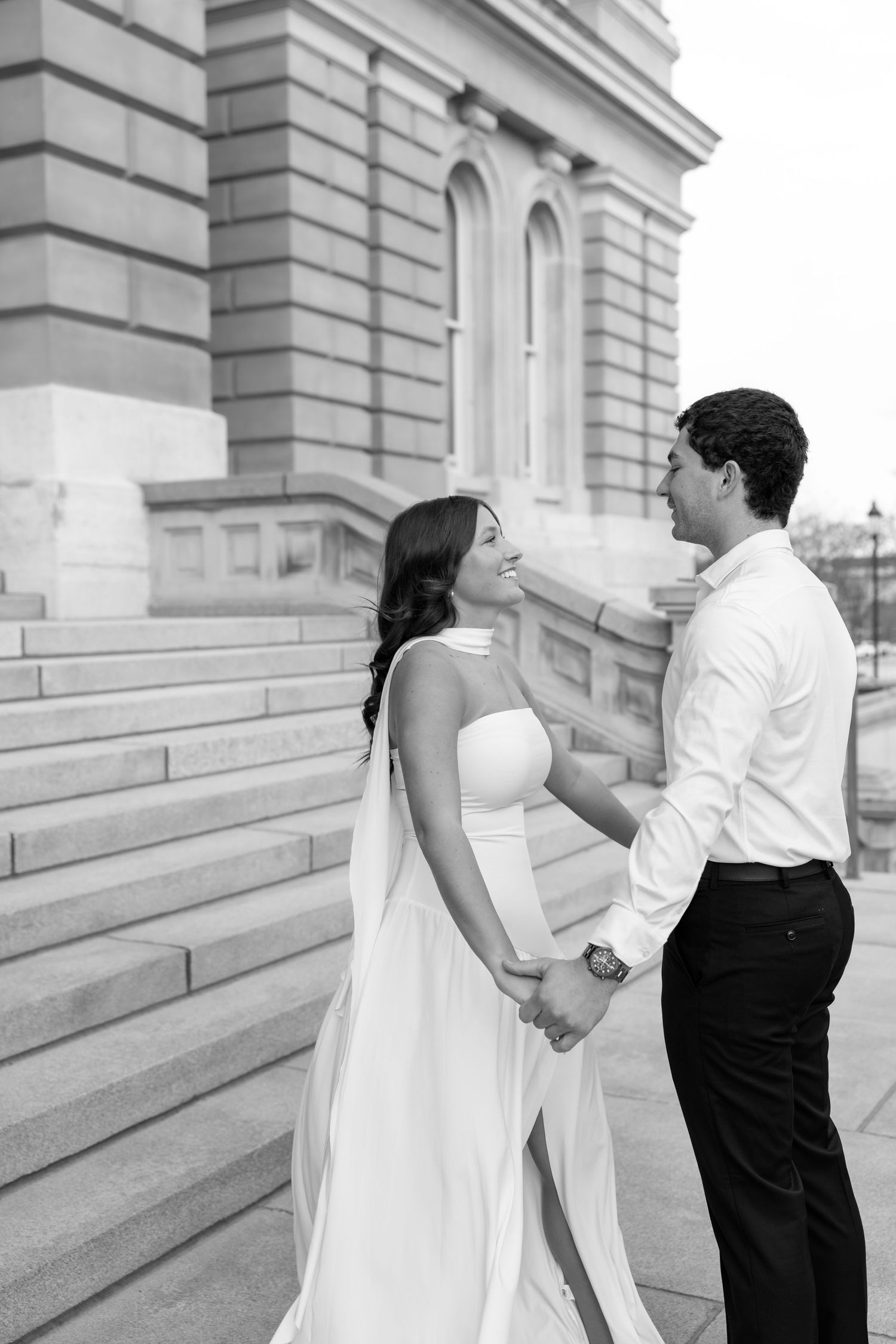 Black and white portrait of bride and groom holding hands and gazing at each other on building steps.