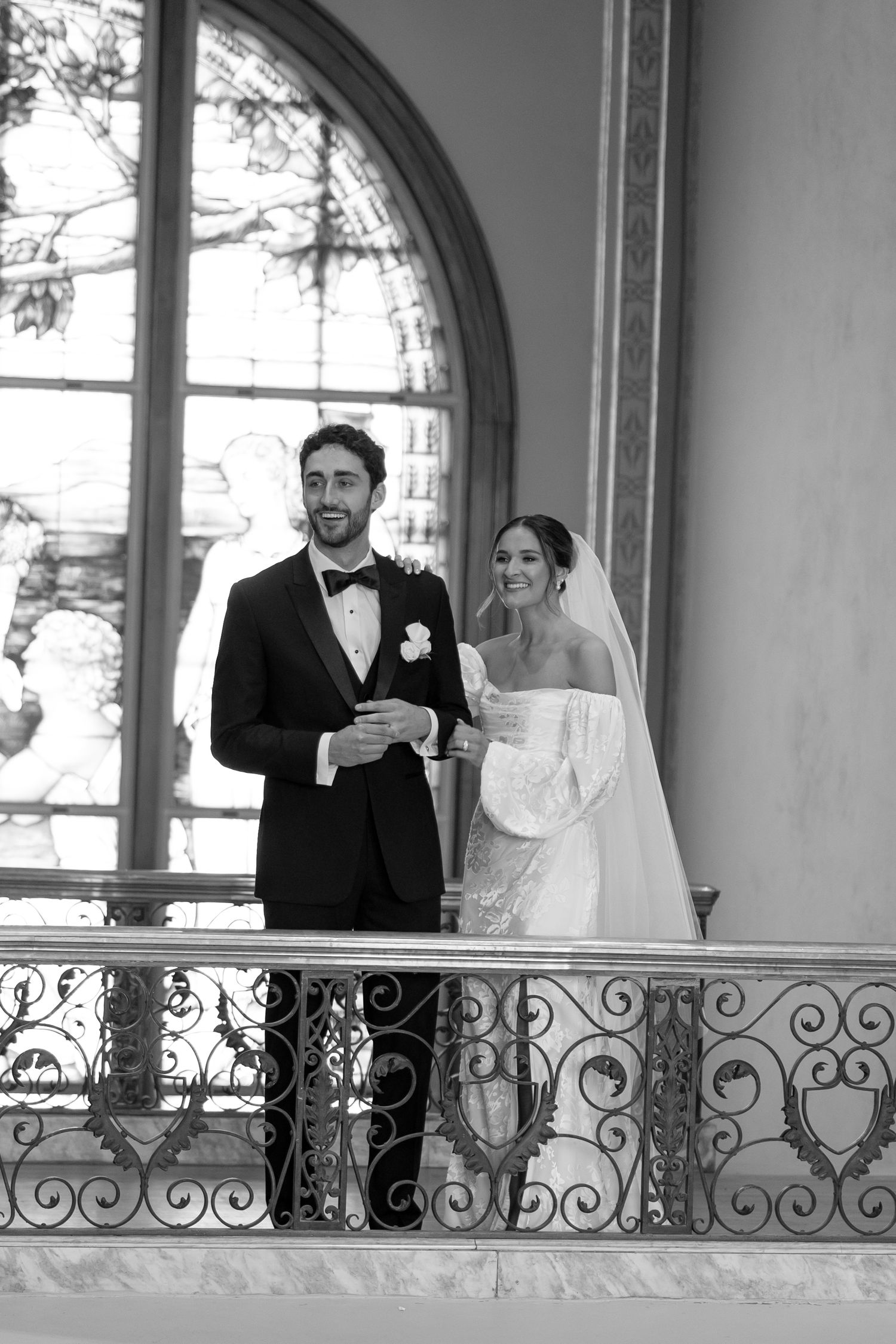 Bride and groom smiling together on ornate iron balcony with stained glass window in black and white.