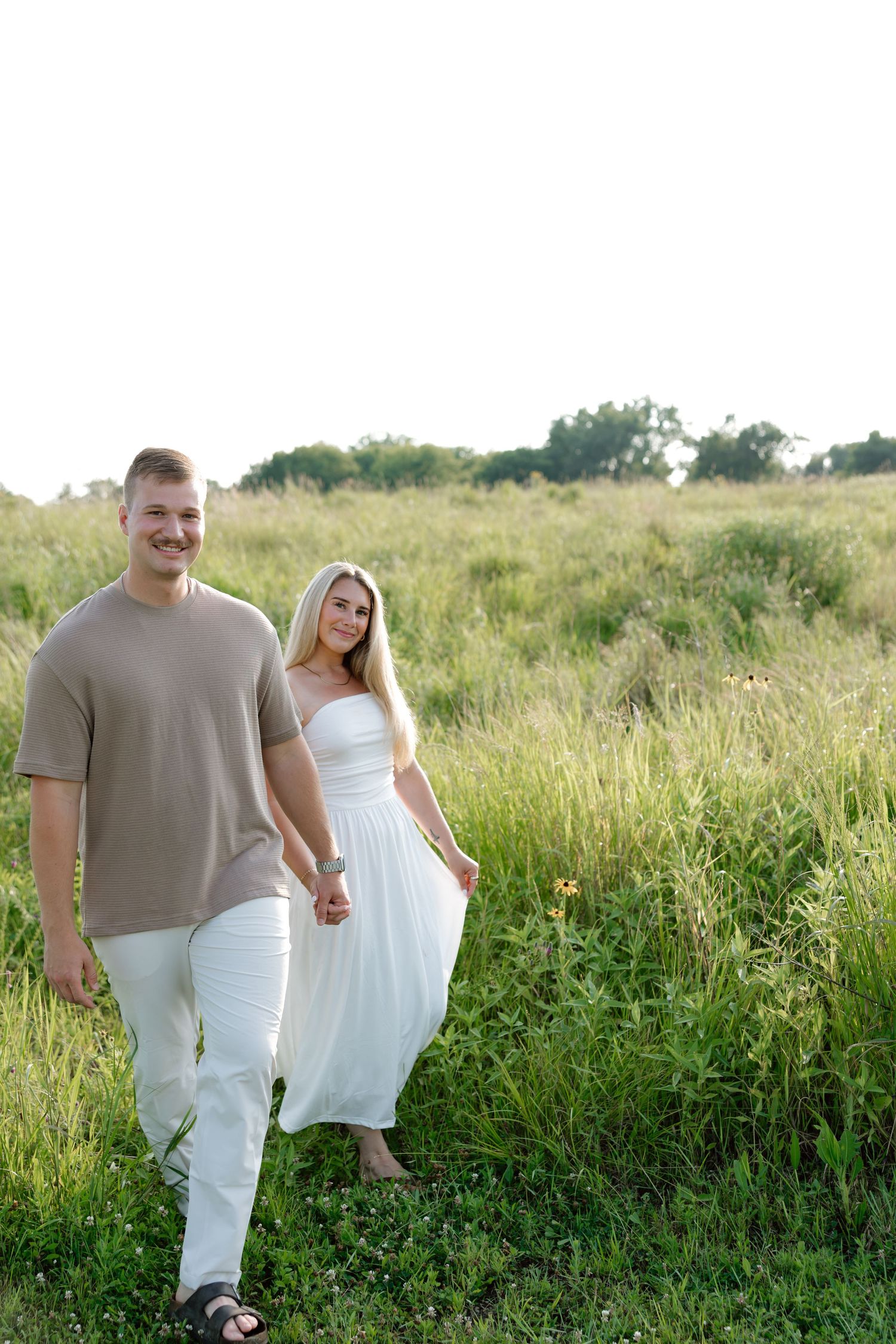 A smiling couple holding hands in a green meadow, man in tan shirt and white pants, woman in white dress.
