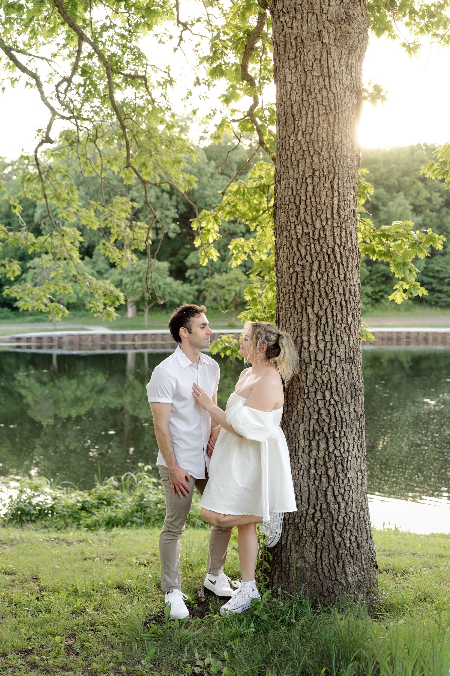 Couple in white outfits standing by a large tree near a pond in a lush green park during golden hour.