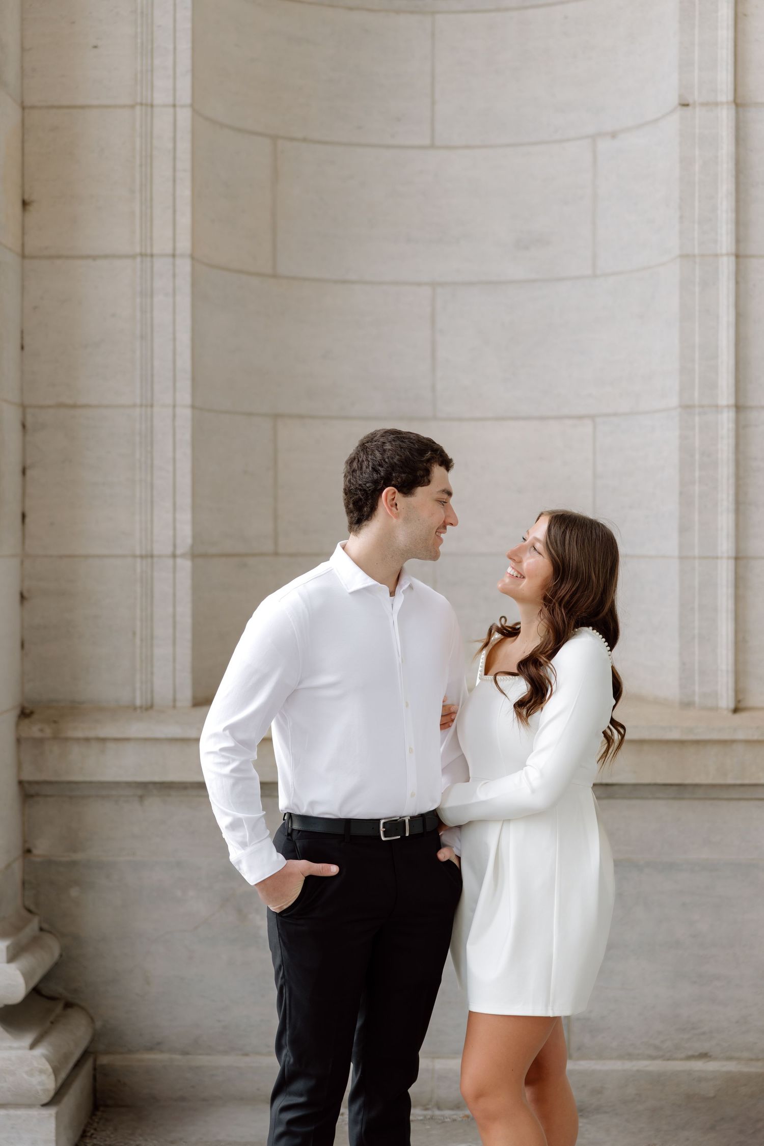 A couple in white outfits smiles at each other while standing beside large stone columns outdoors.