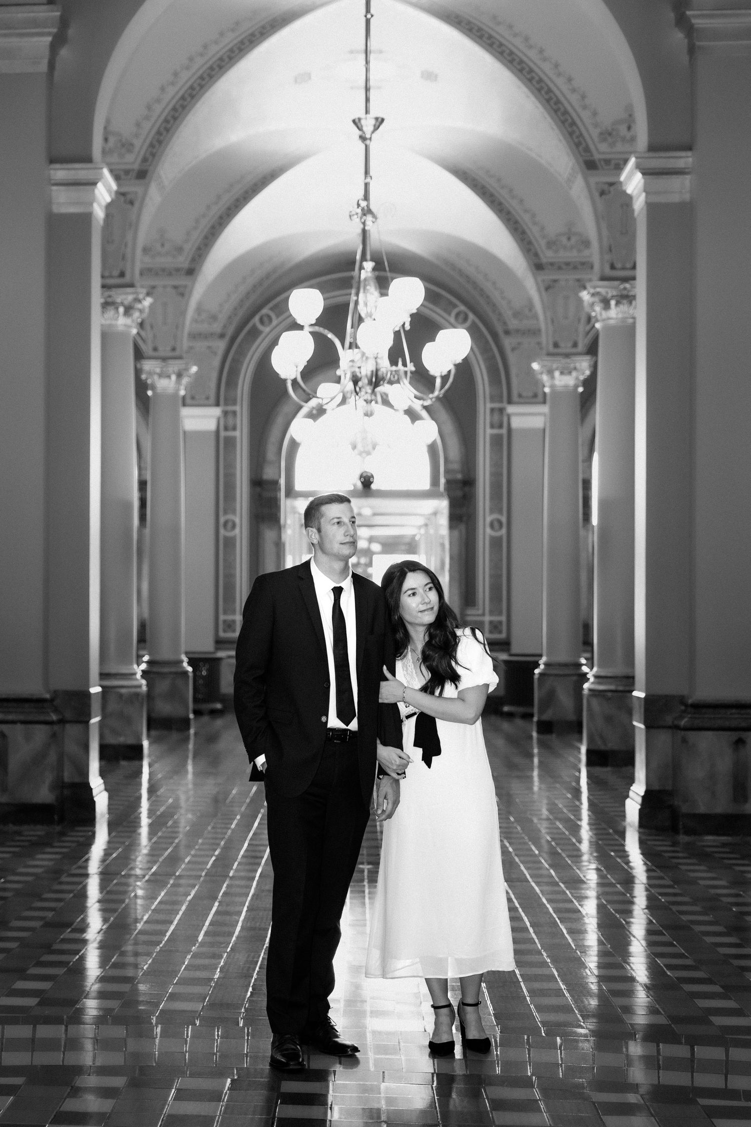 Elegant black and white photo of a couple standing in a grand hallway with arched ceilings and a chandelier.