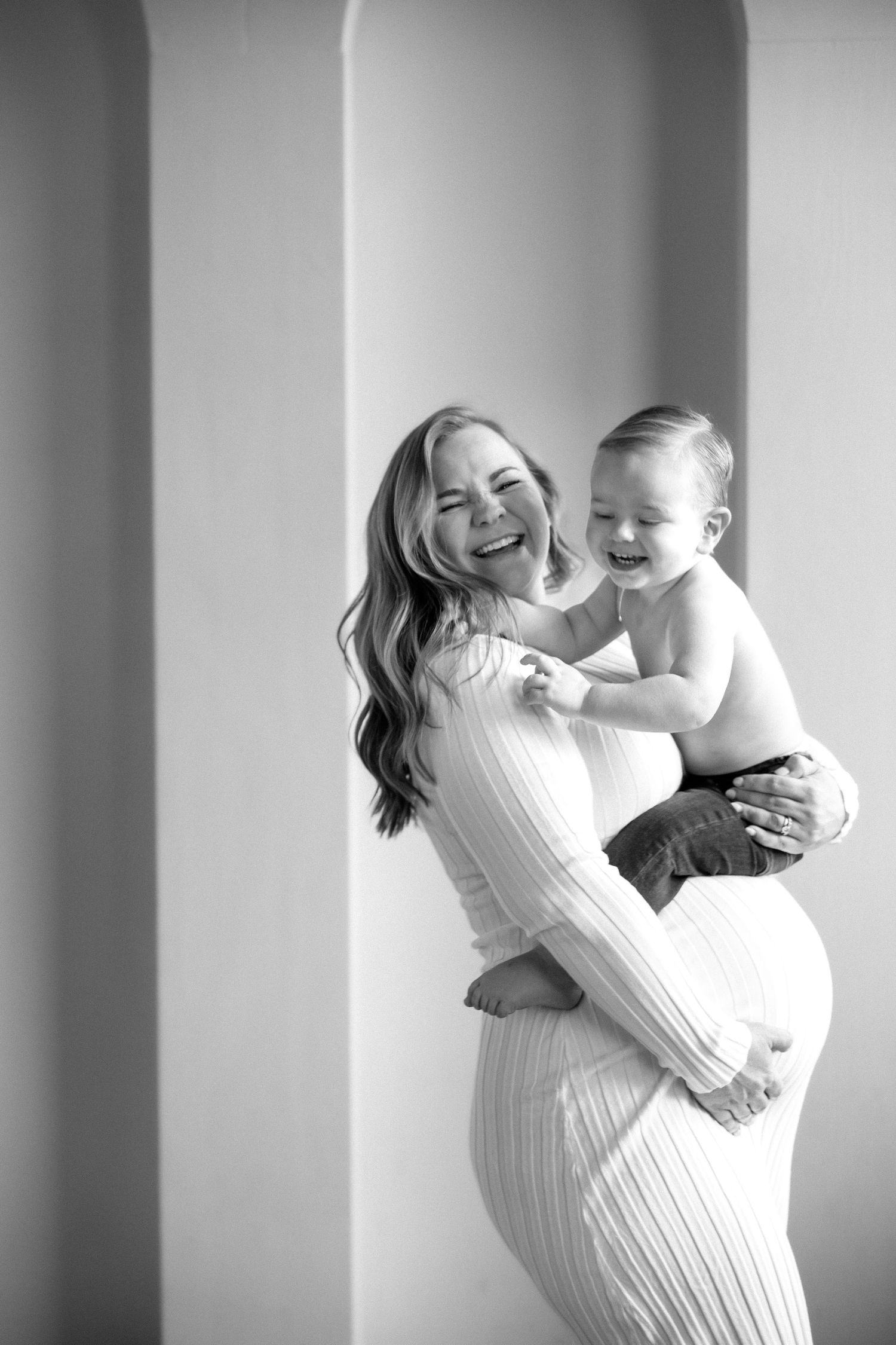 Black and white photo of a laughing pregnant woman in white dress holding her giggling toddler son.