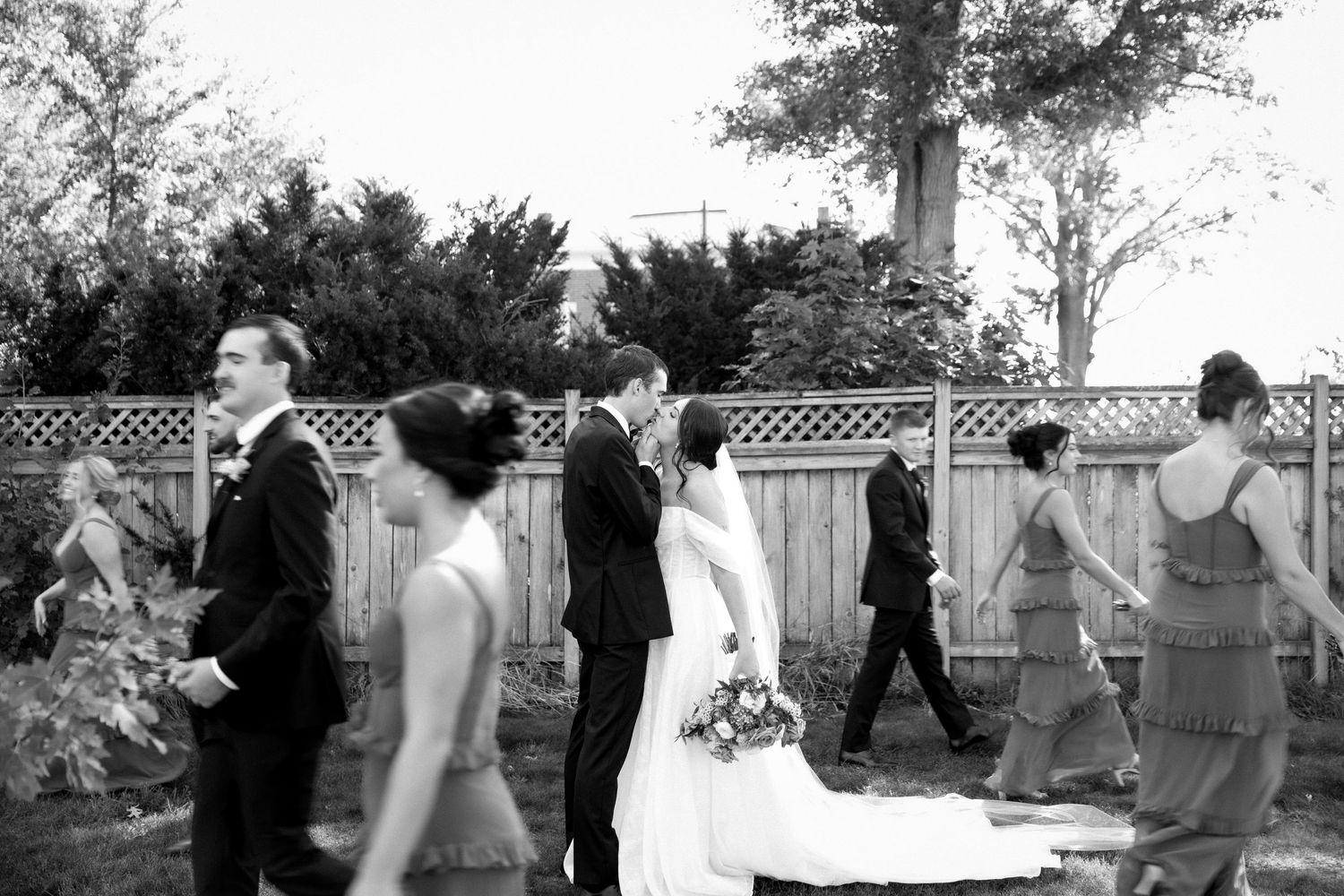 Black and white photo of wedding party walking outdoors, bride and groom kissing while bridesmaids and groomsmen pass by.