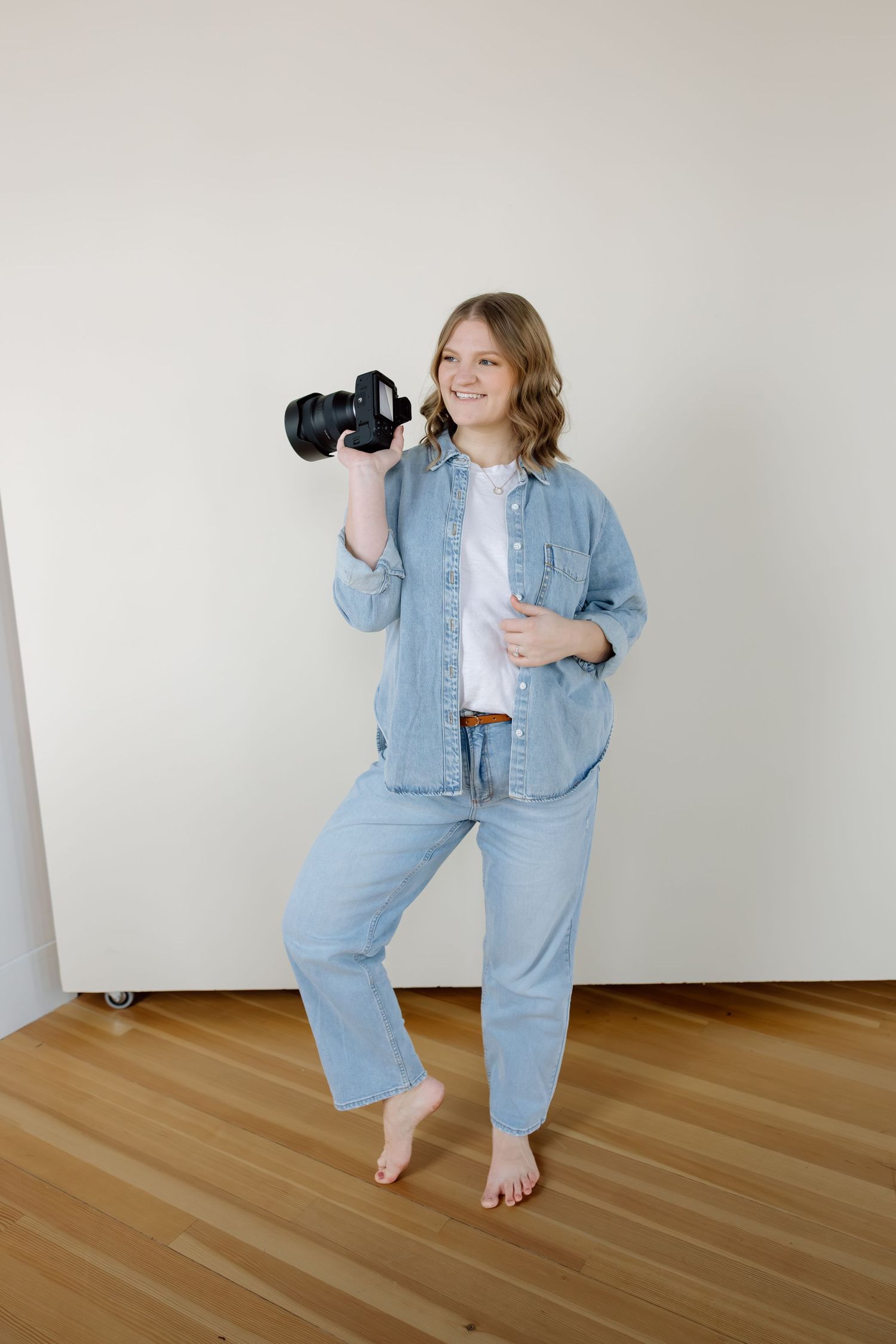 A cheerful female photographer in denim outfit holds a professional camera in a bright studio space.