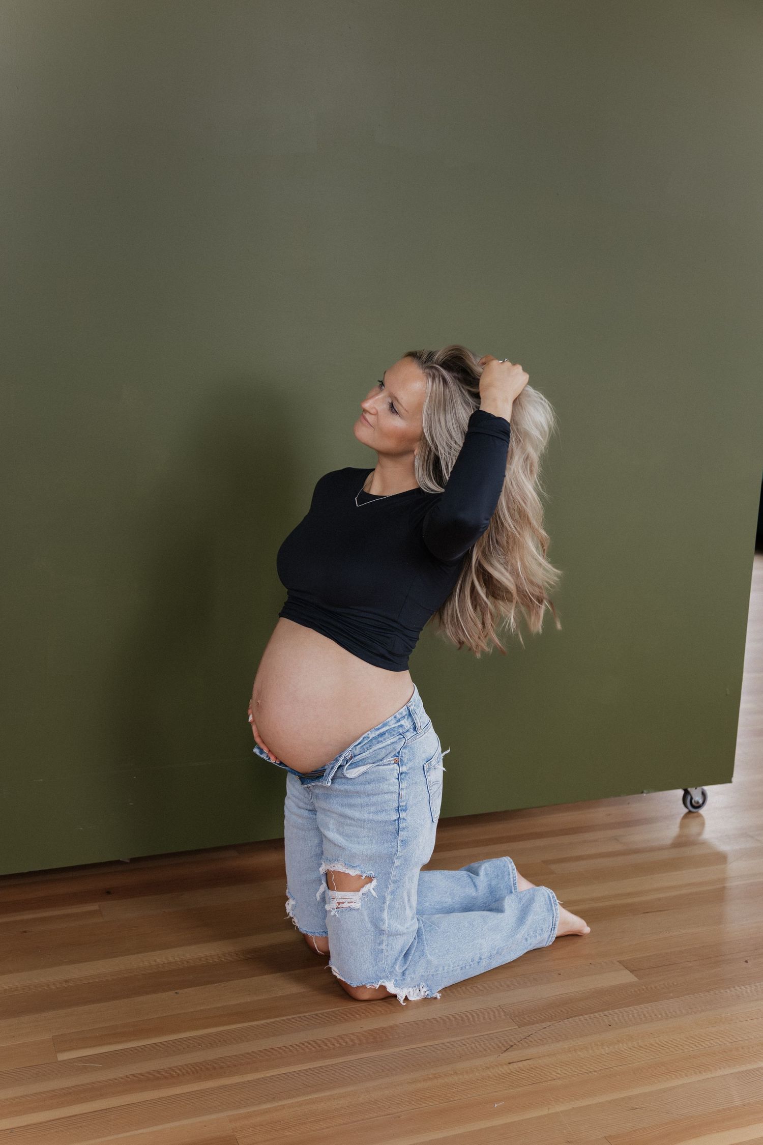 Pregnant woman kneeling on floor against green wall, smiling while lifting her long blonde hair up.