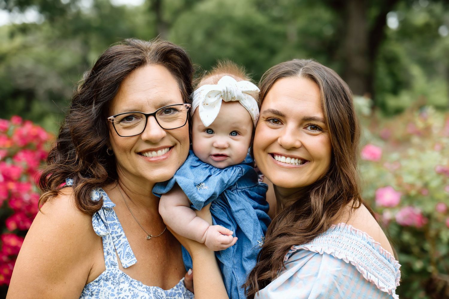 Two women smiling with happy baby girl in blue dress and white bow in a garden.