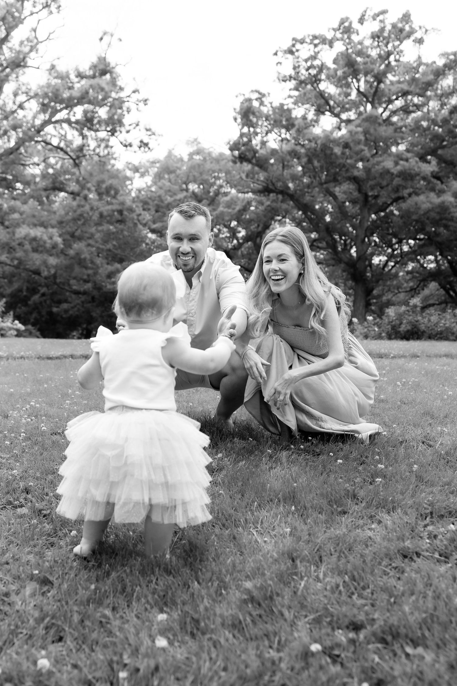 Black and white photo of parents crouching on grass as their toddler daughter in tutu walks toward them.