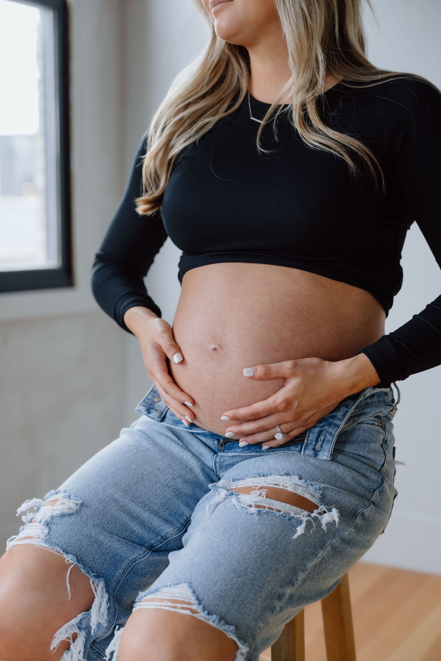 Close-up of pregnant woman in black crop top gently holding her bare baby bump while seated on a stool.