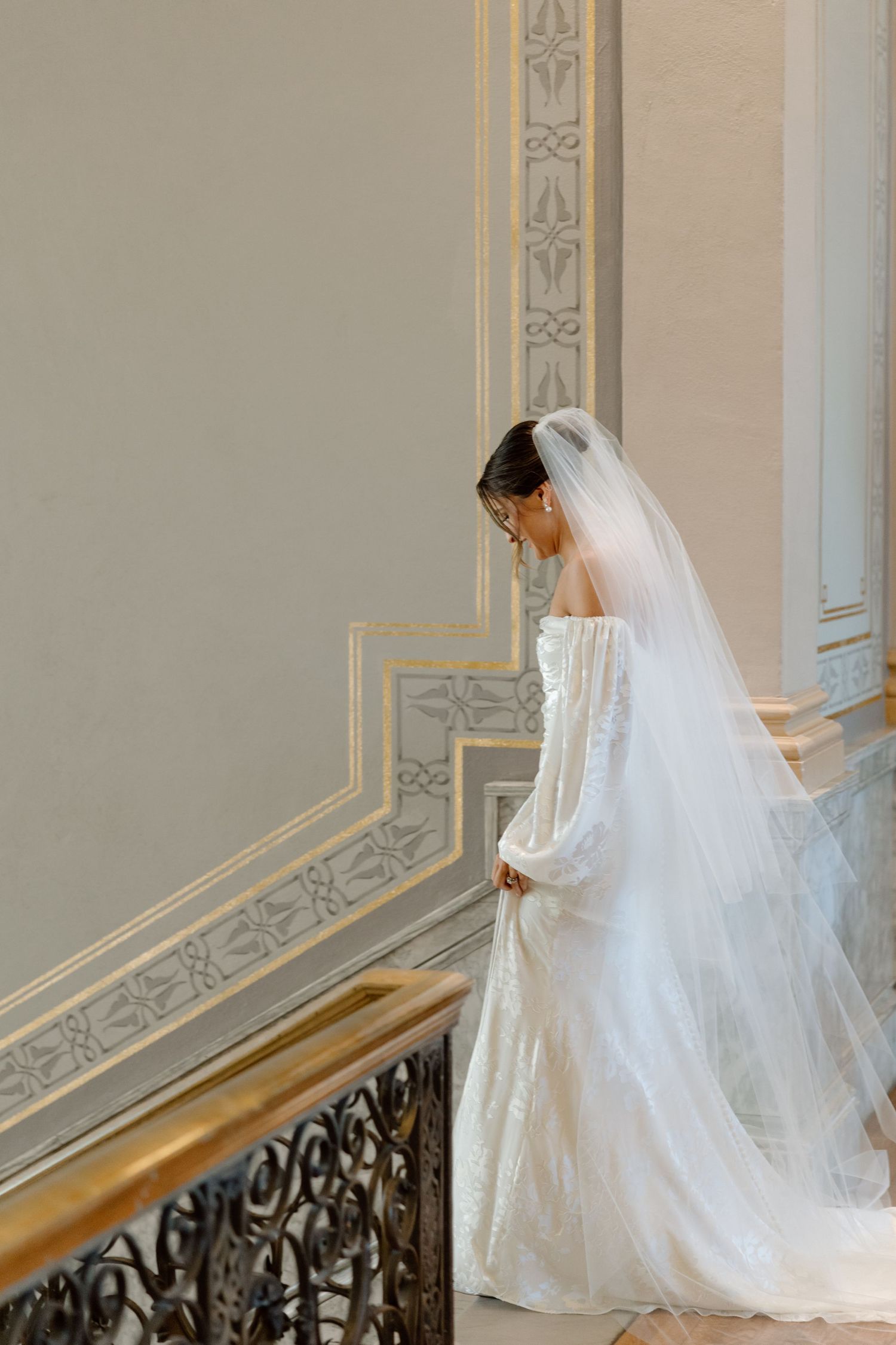 Bride in white off-shoulder gown and veil stands gracefully on ornate staircase in elegant venue.