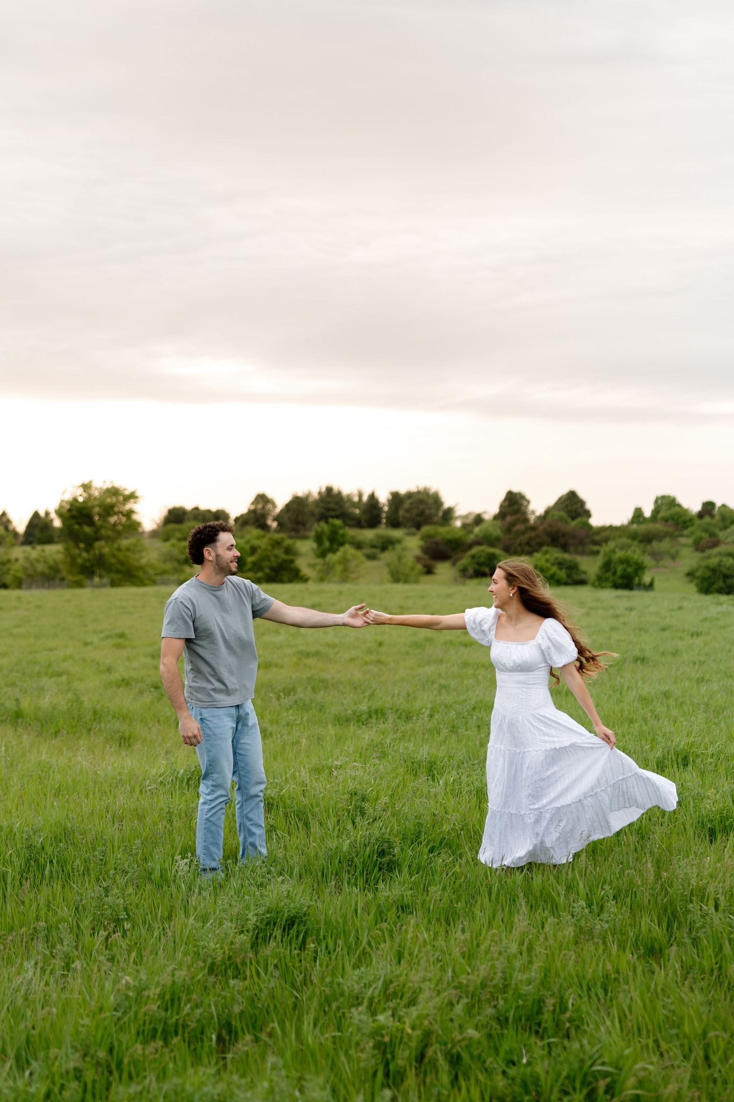 Couple holding hands and dancing in a lush green field, woman in white dress, man in gray shirt and jeans.